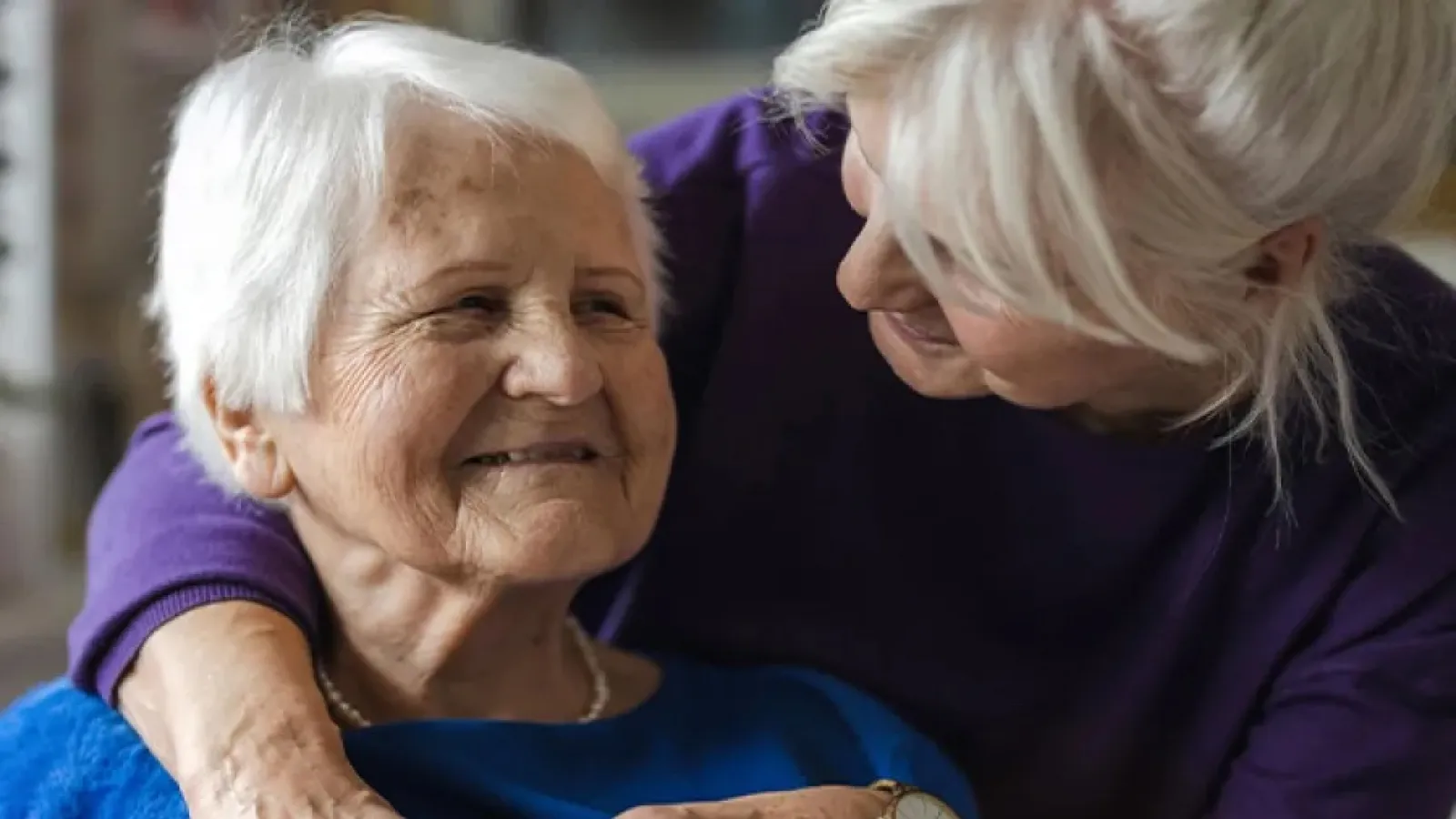 Elderly woman smiling warmly as another woman embraces her from behind showing care and affection.