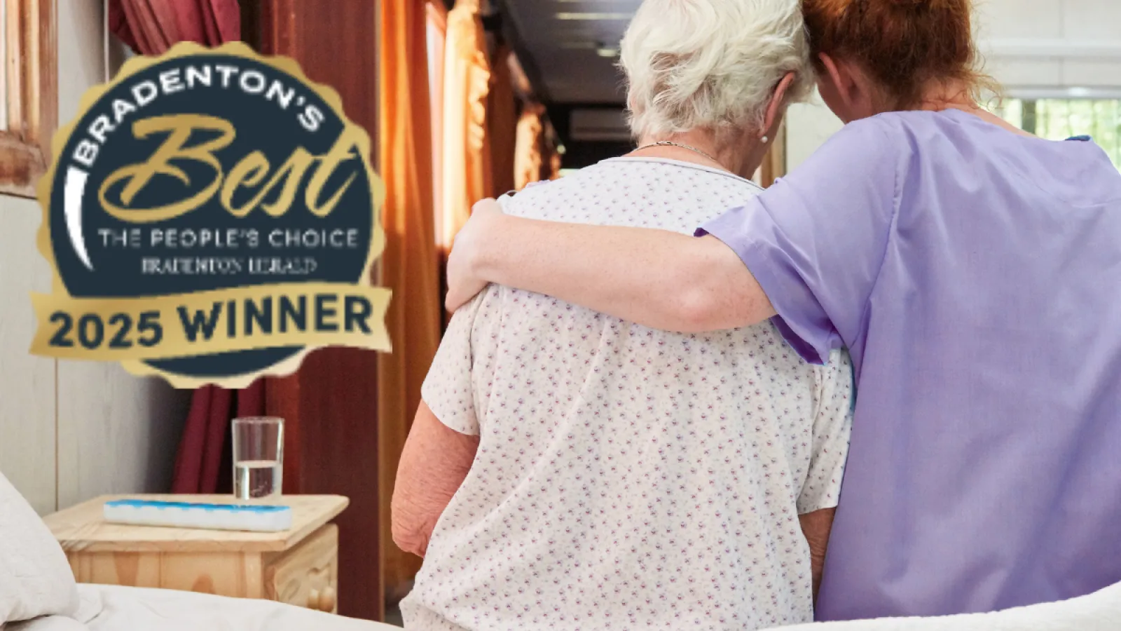 Caregiver in purple scrubs embraces elderly woman sitting on bed in cozy room with 2025 winner badge.
