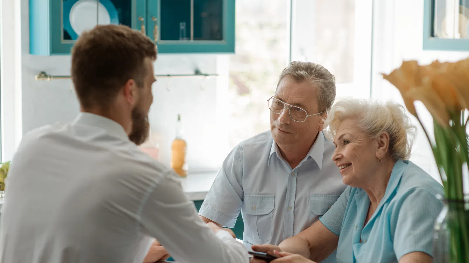 Senior couple consulting with a professional in a bright kitchen, discussing documents and holding a smartphone.