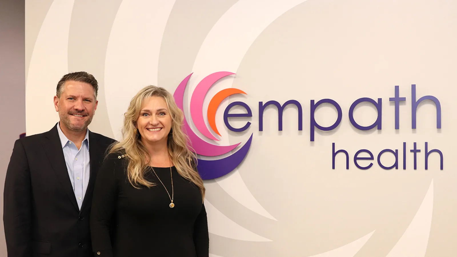 Two professionals standing and smiling beside Empath Health logo on a beige wall in office setting