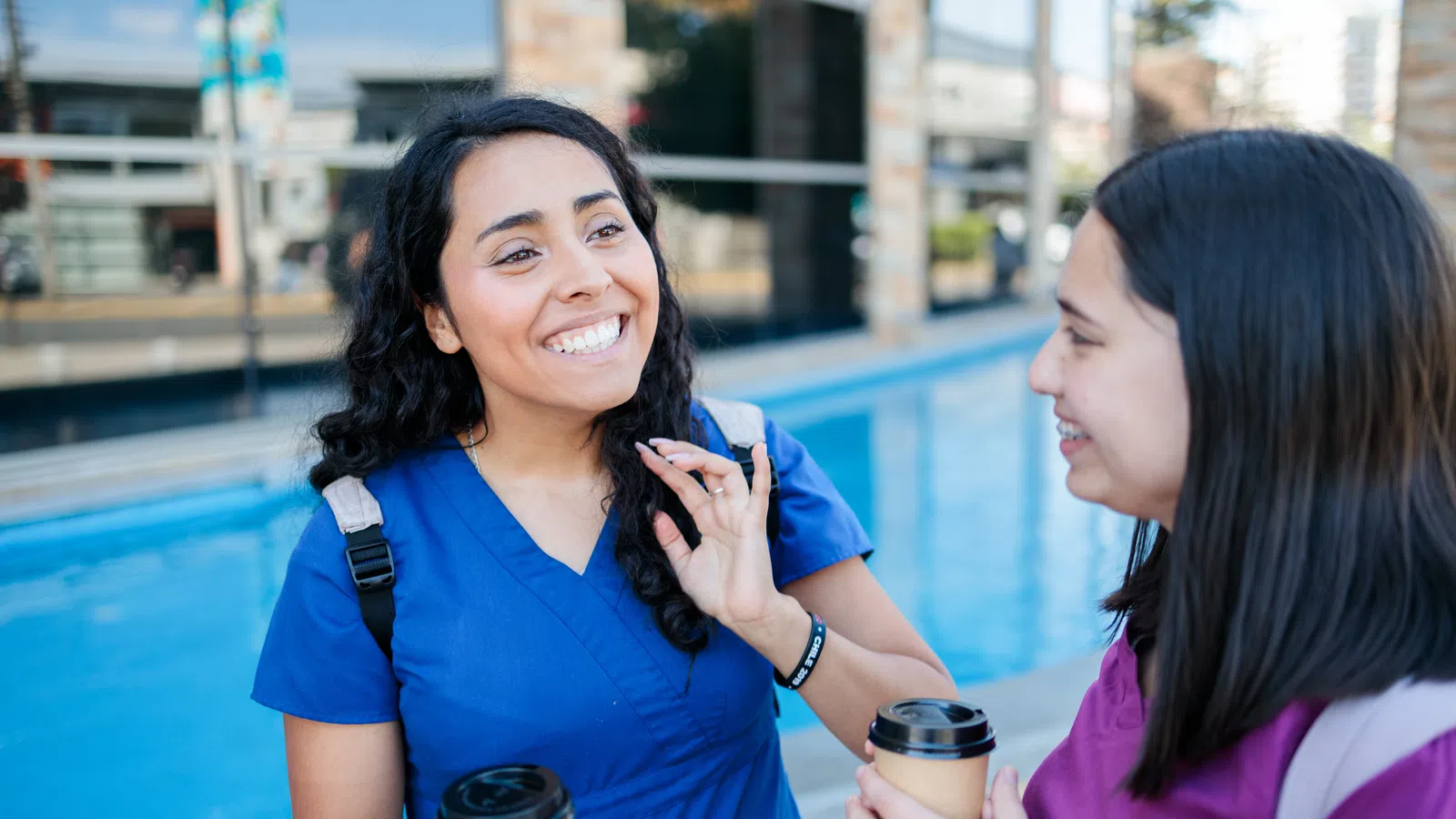 Two smiling women in scrubs holding coffee cups near a poolside setting.