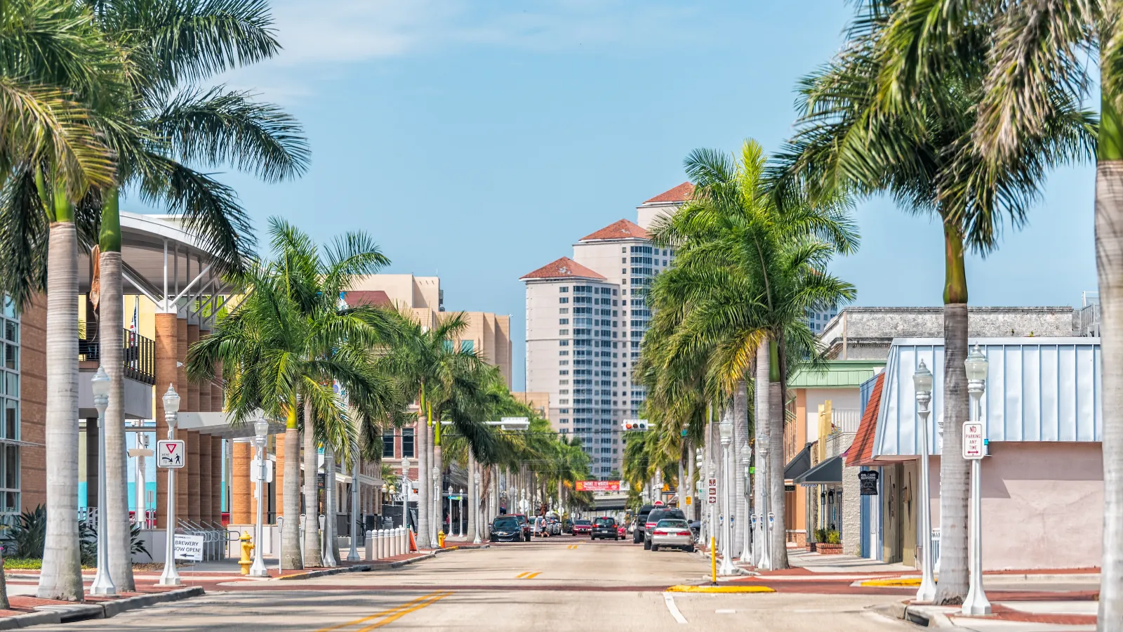 Downtown street lined with tall palm trees, low-rise buildings, and high-rise towers under a blue sky.