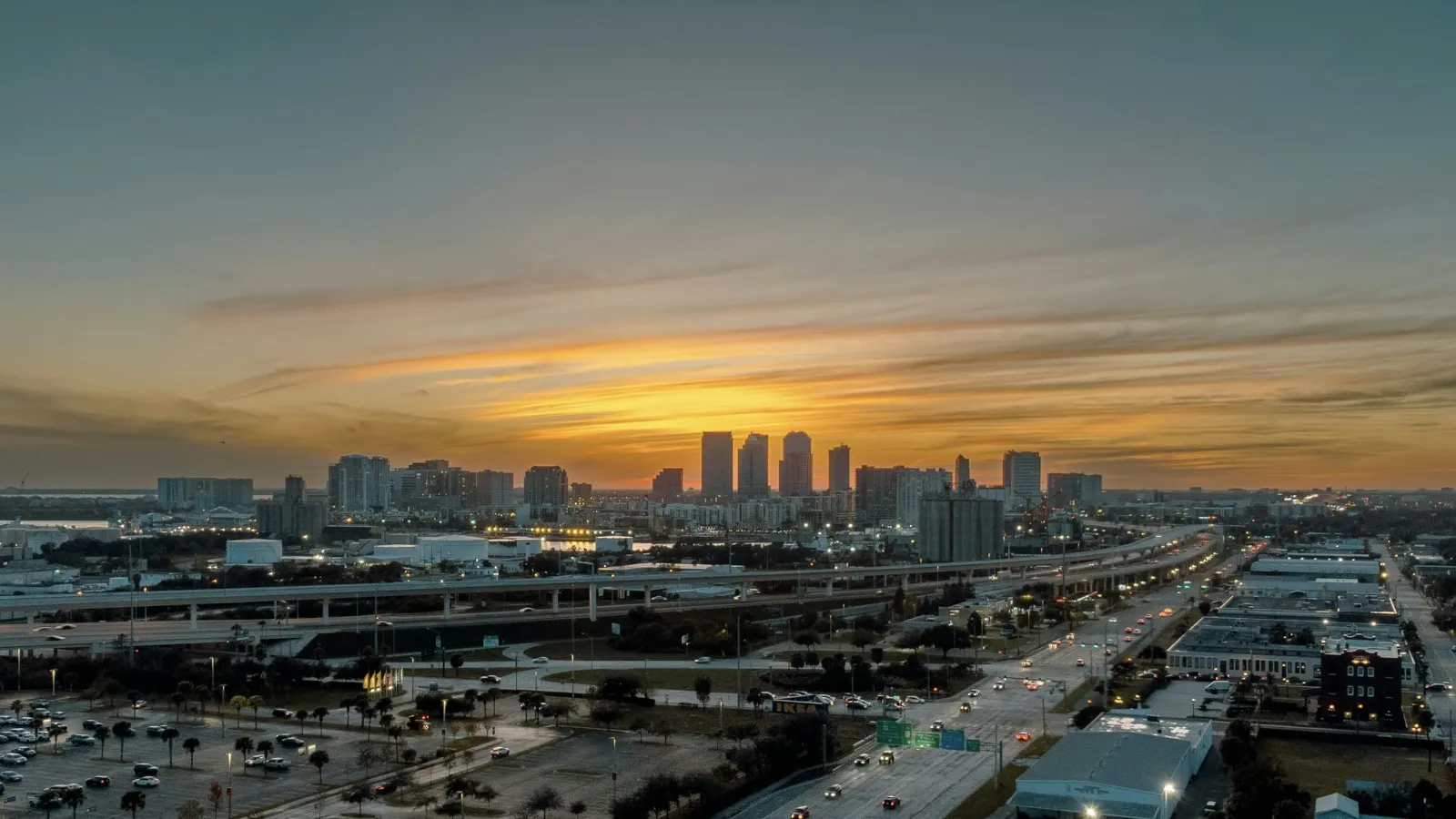 Sunset over a city skyline with tall buildings and busy highway traffic creating an urban evening scene.