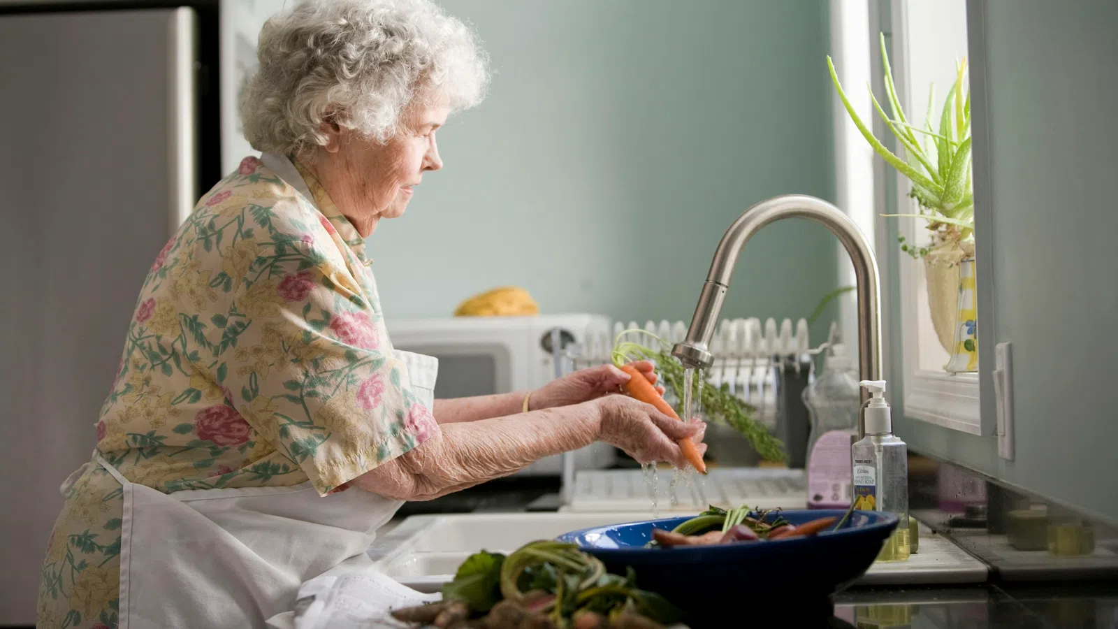 Elderly woman washing fresh carrots under kitchen sink faucet preparing vegetables for cooking.