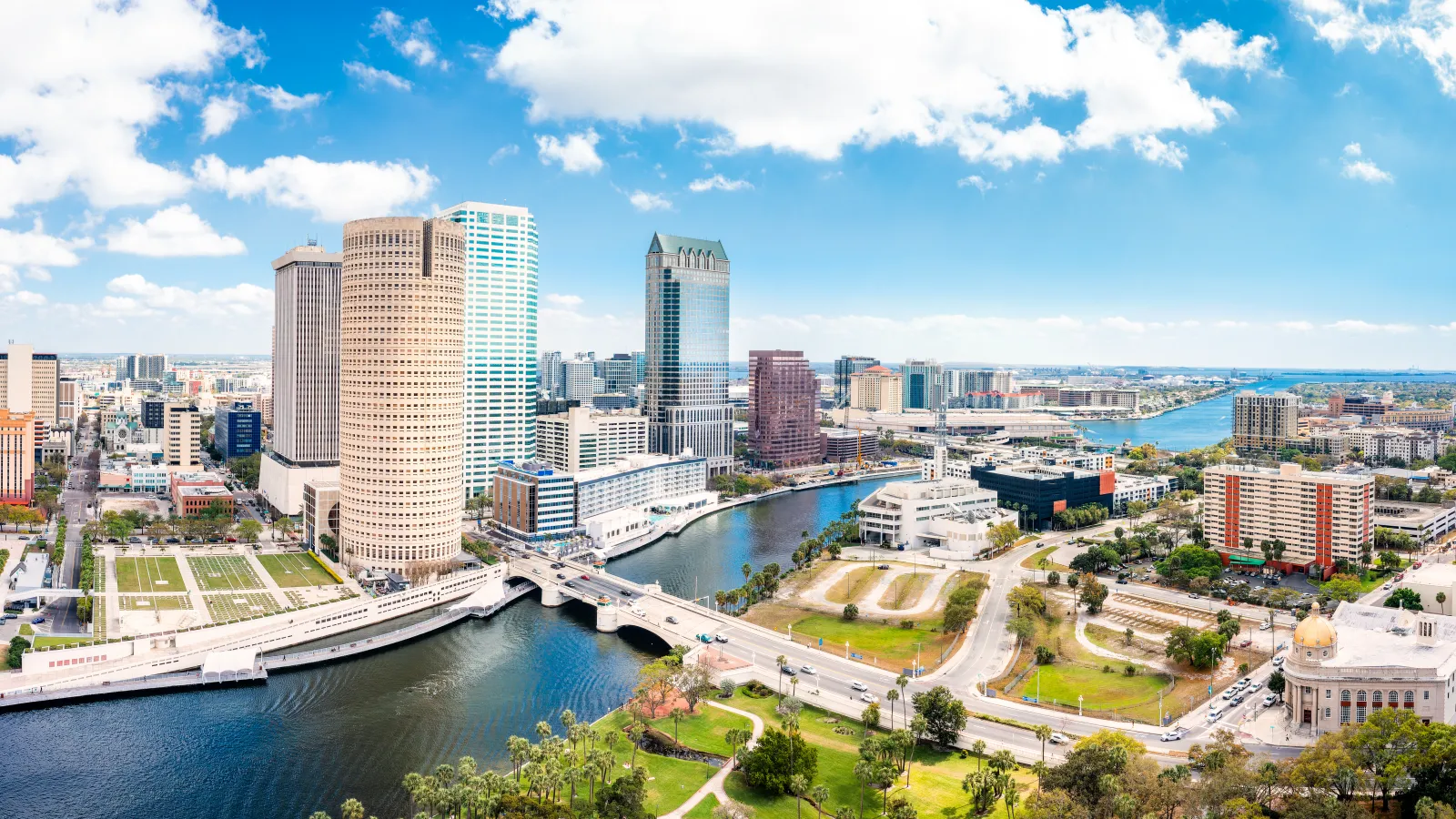 Aerial view of Tampa skyline with Hillsborough River, urban buildings, bridges, and blue sky with clouds.