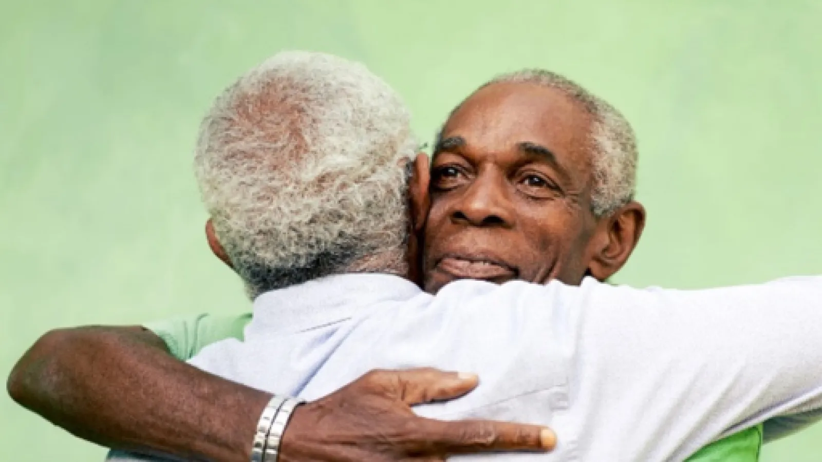 Two older men embracing in a warm hug against a soft green background, showing connection and friendship.