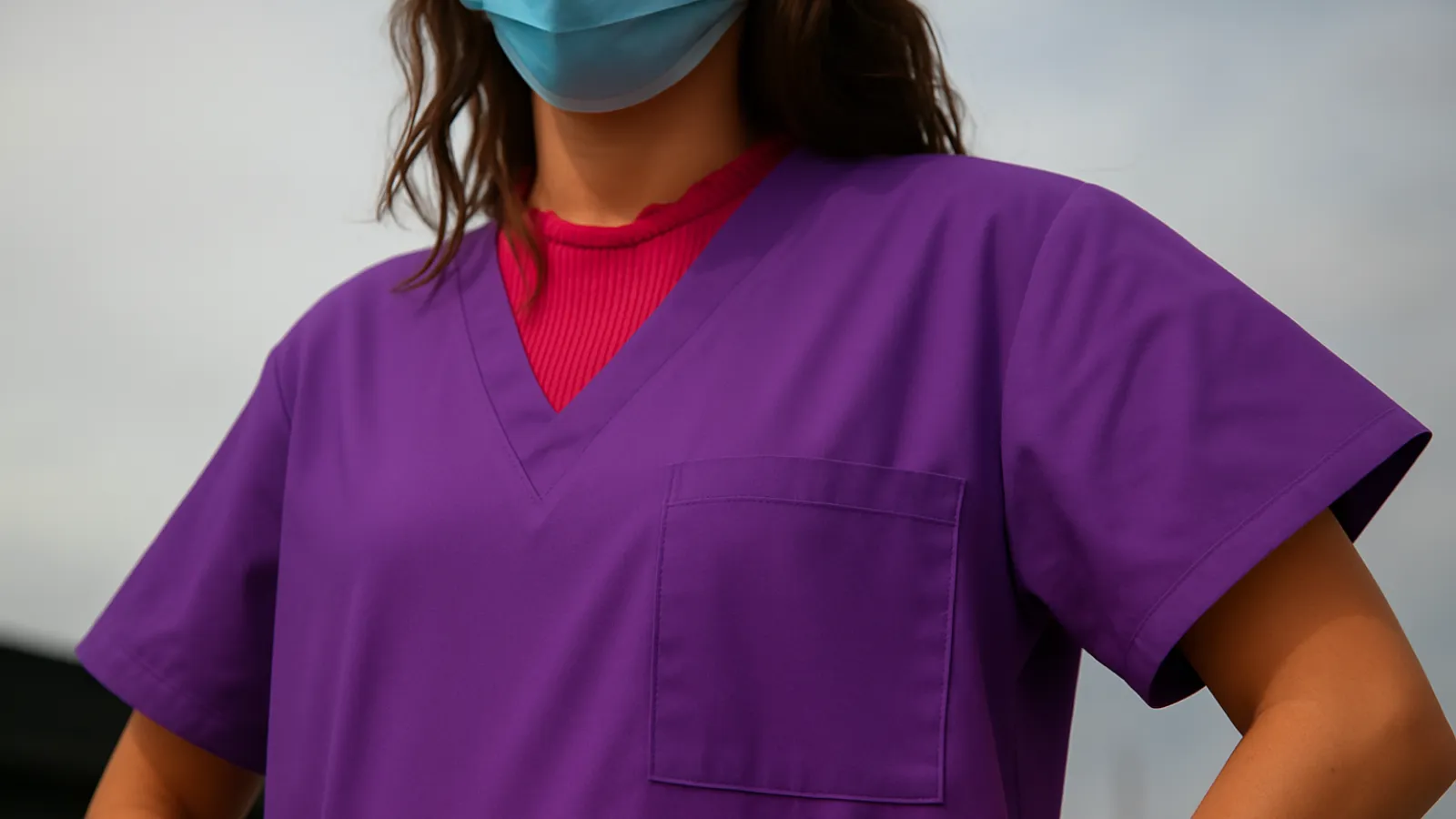 Healthcare worker wearing purple scrubs and a blue face mask standing outdoors with hands on hips.