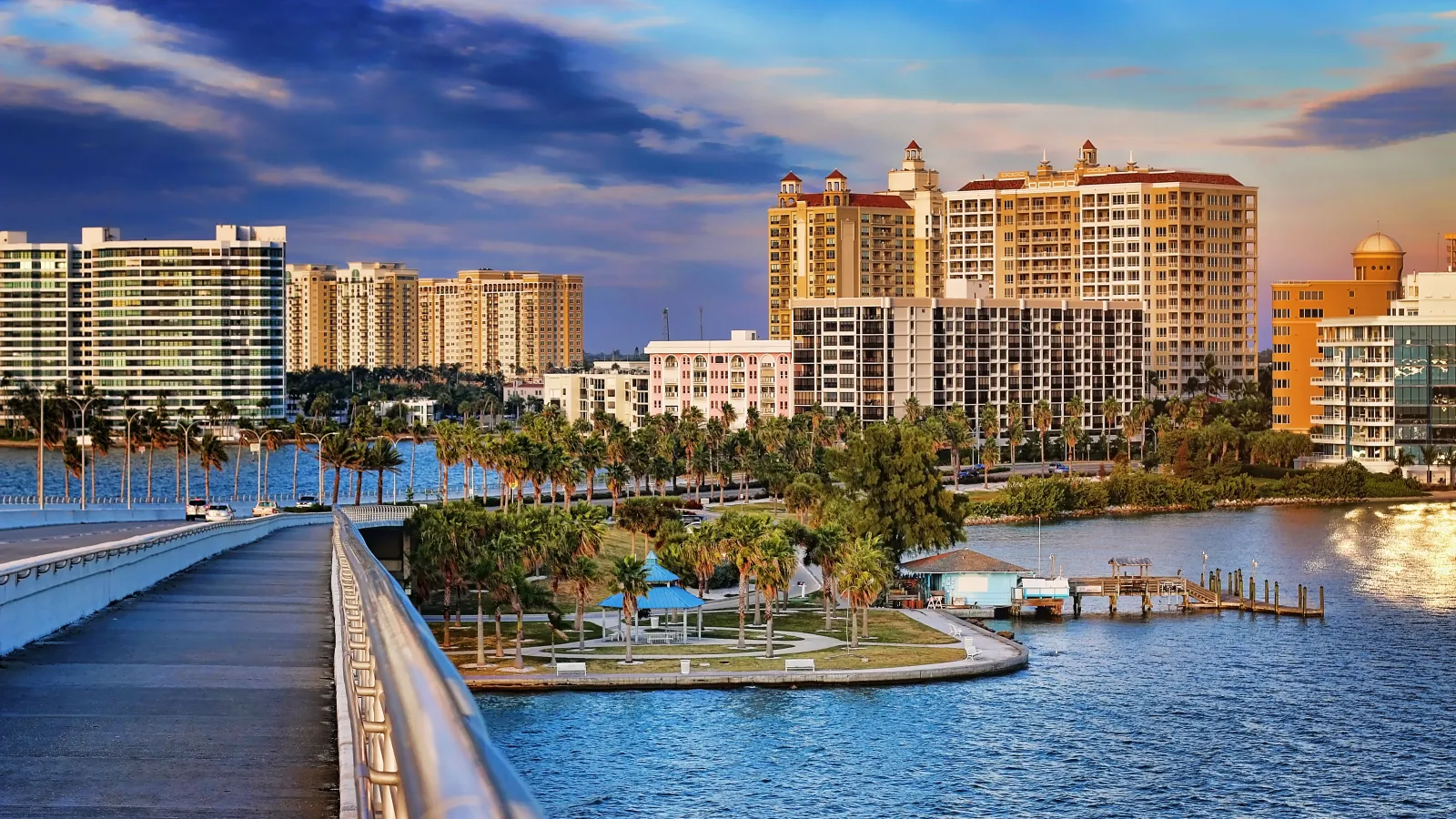 Waterfront cityscape with modern high-rise buildings, palm trees, and a bridge at sunset under partly cloudy sky
