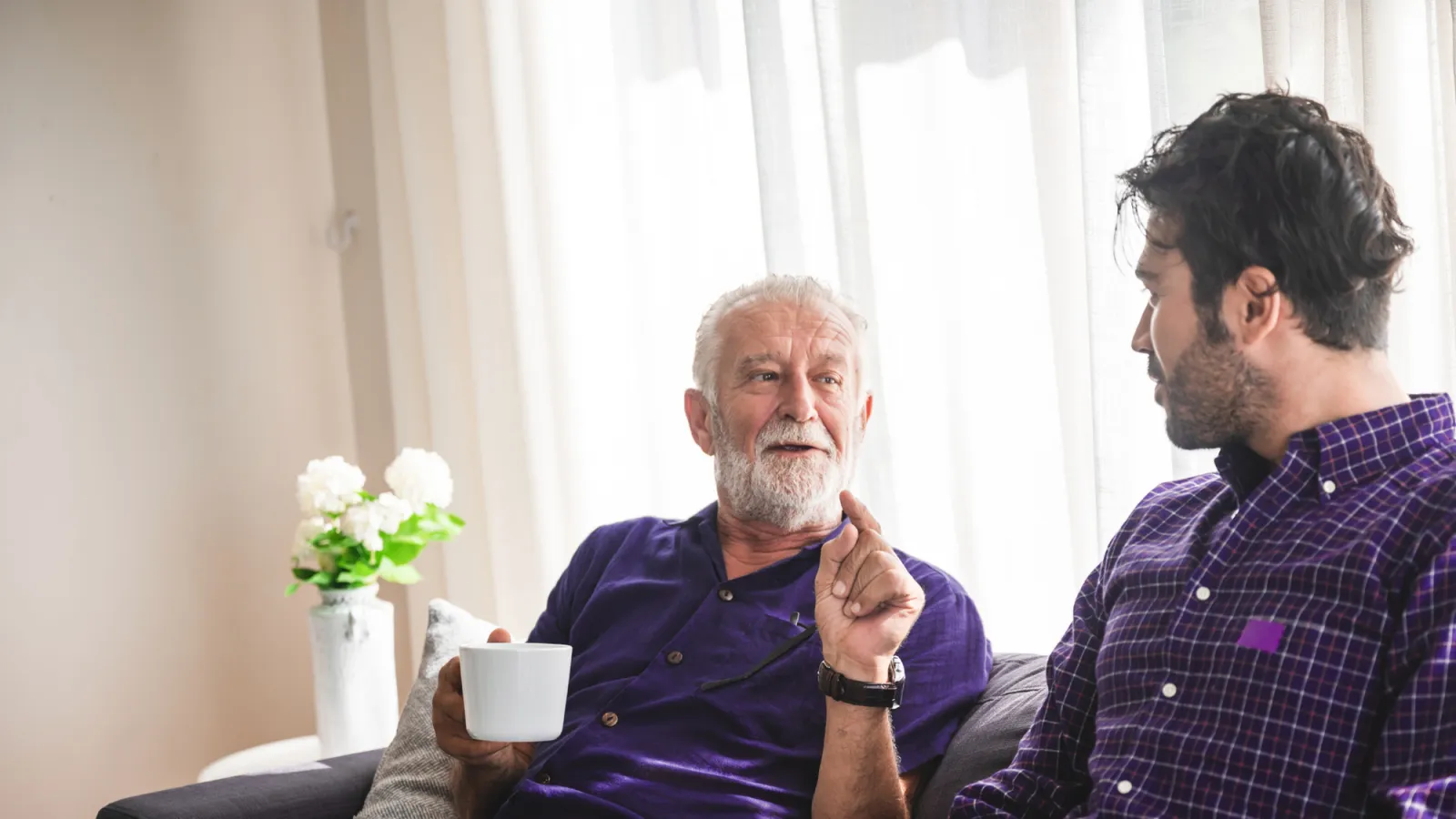 Elderly man and younger man sitting on couch drinking coffee and having a conversation in a bright room