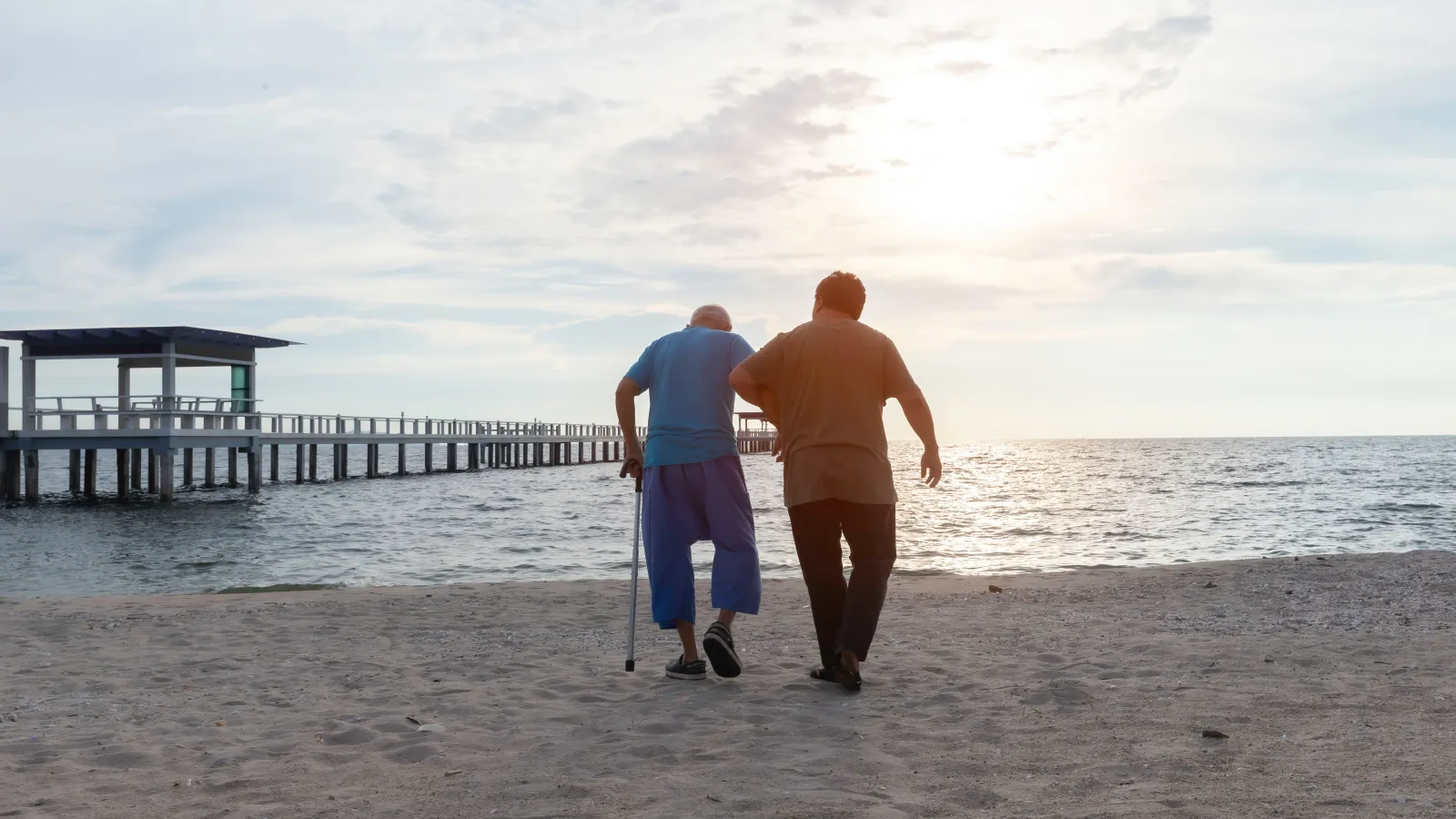 Two men walking on the beach at sunset, one using a cane for support, next to a wooden pier extending into the sea.