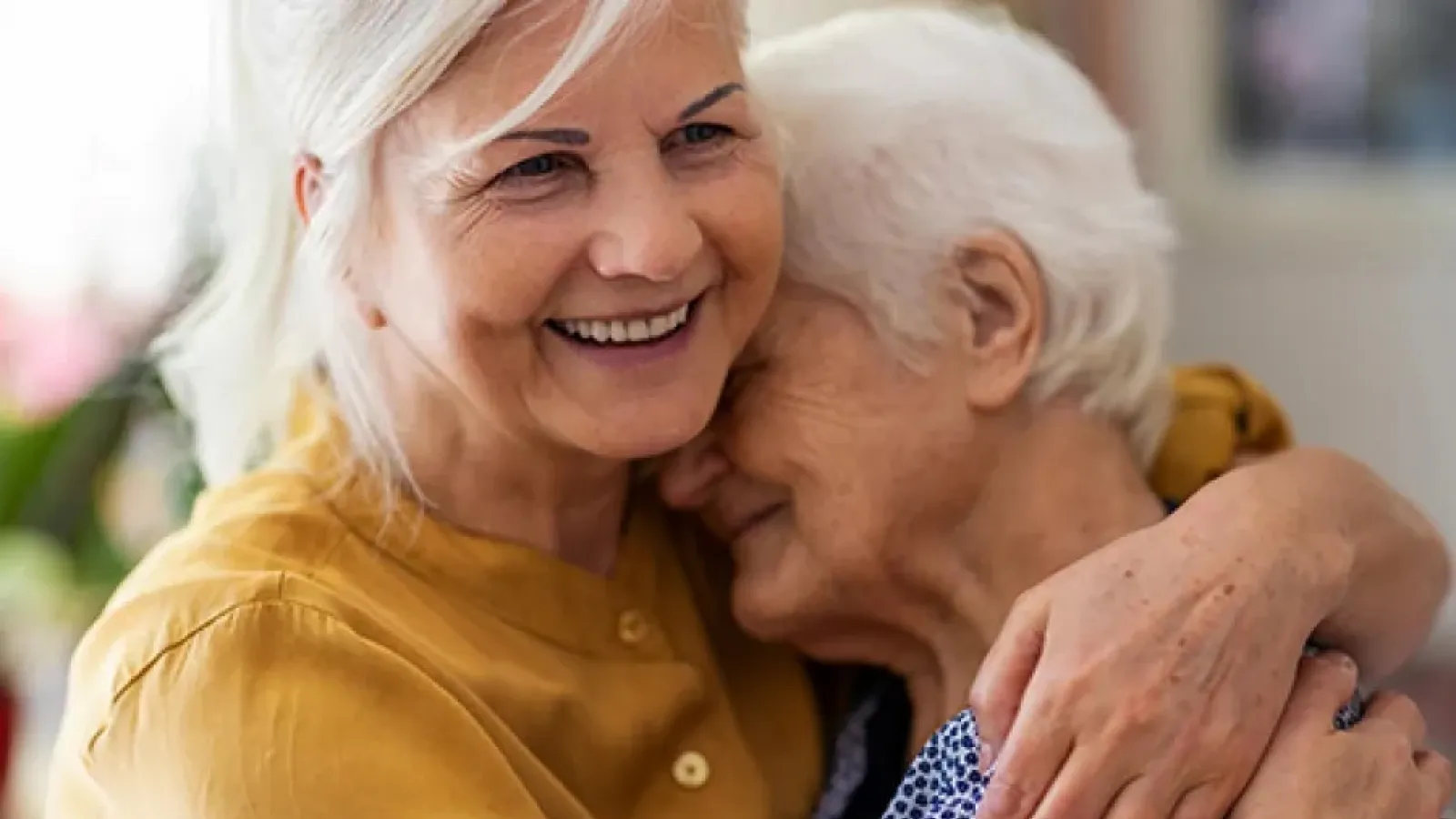 Smiling middle-aged woman warmly hugging elderly woman in a close, loving embrace indoors