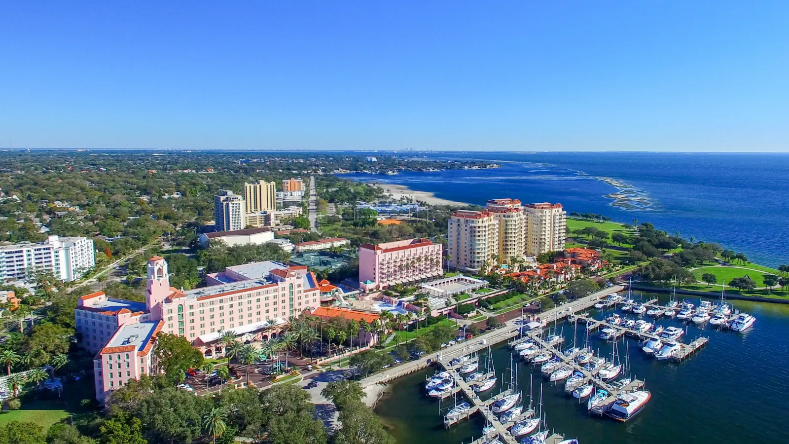 Aerial view of a coastal city with yachts docked at the marina, beachfront hotels, and clear blue ocean under a sunny sky