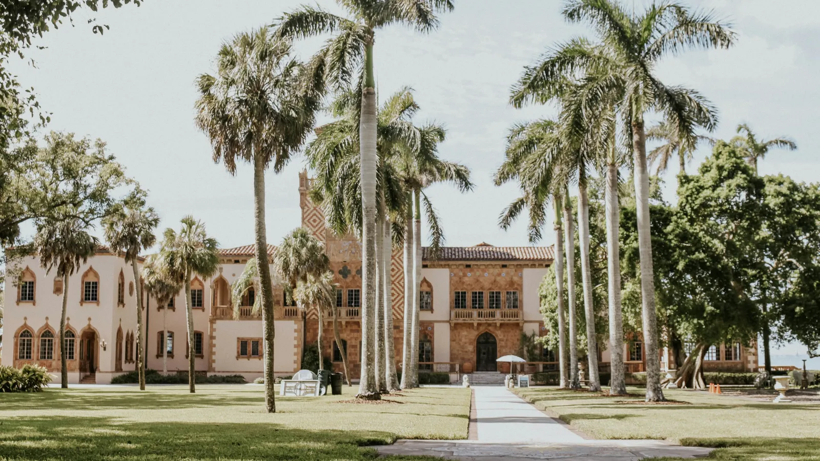 Historic Mediterranean-style building with tall palm trees lining a walkway surrounded by green lawns under a clear sky.