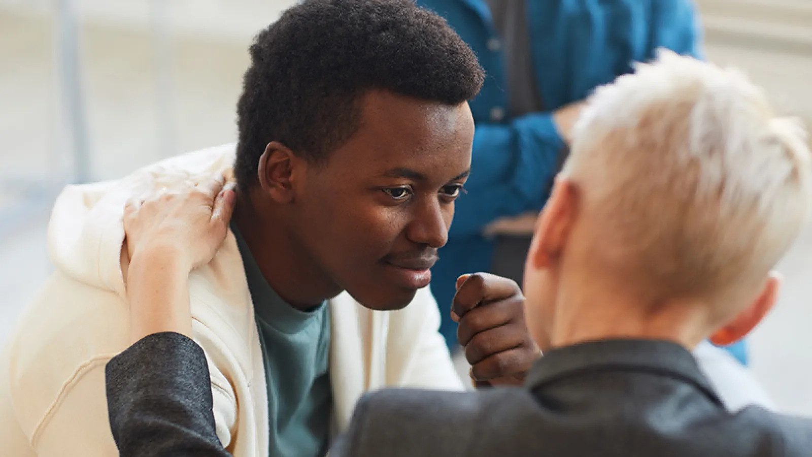 Young man in white hoodie receiving support from a blonde person in gray jacket during a conversation.