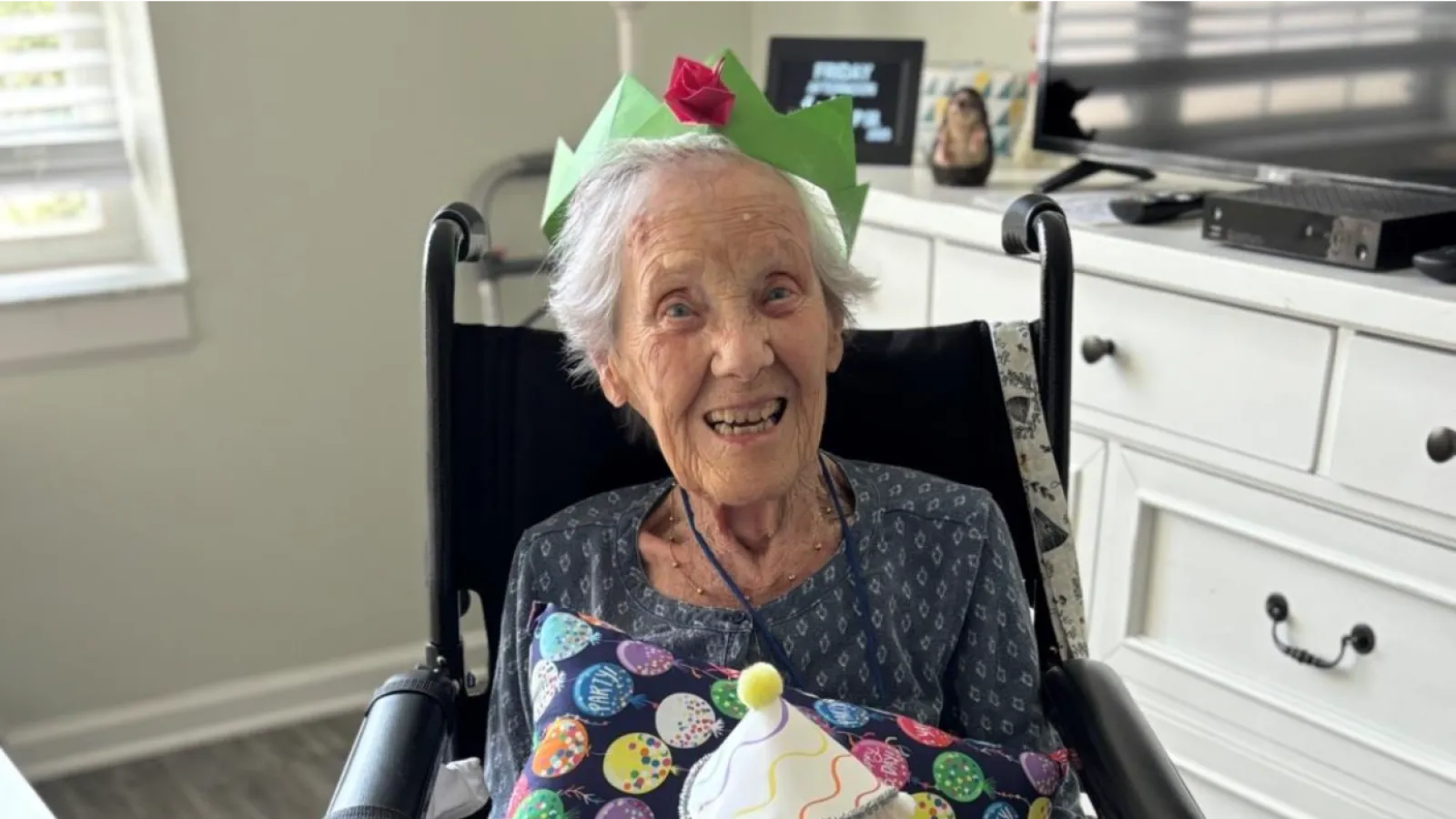 Elderly woman in a wheelchair wearing a green paper crown, smiling and holding a teddy bear with a party hat.