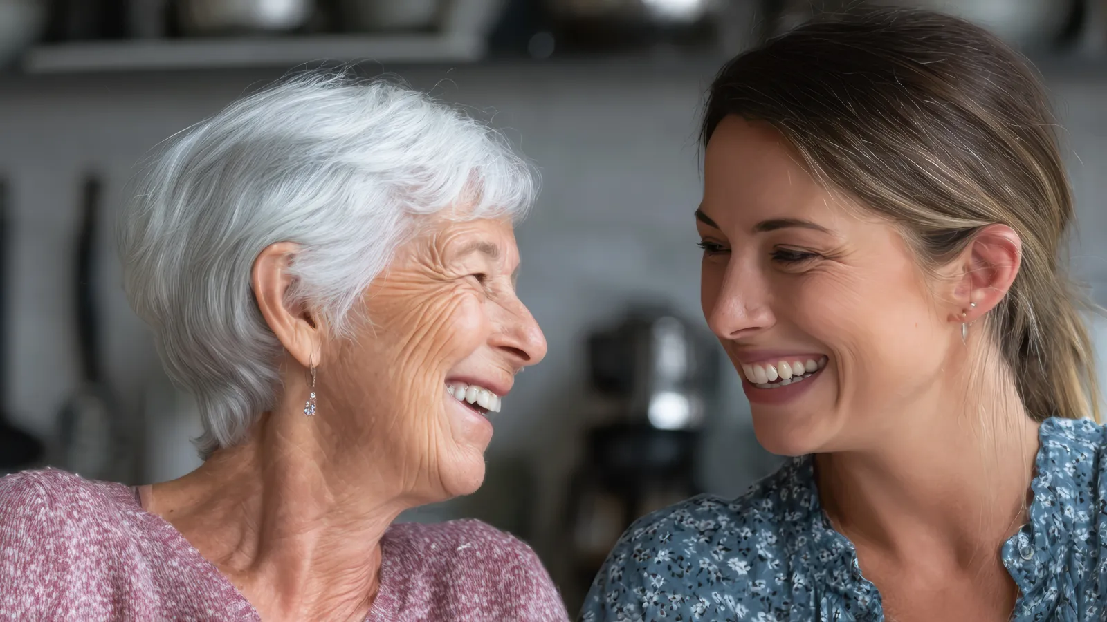 Smiling elderly woman and younger woman sharing a joyful moment together in a cozy kitchen setting.