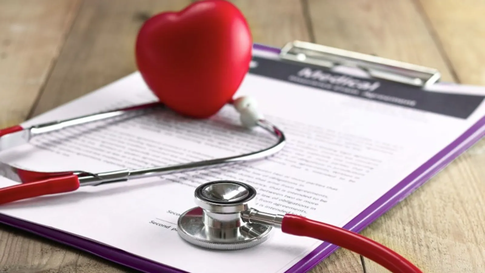 Red stethoscope and heart model on medical documents on a wooden desk representing healthcare.