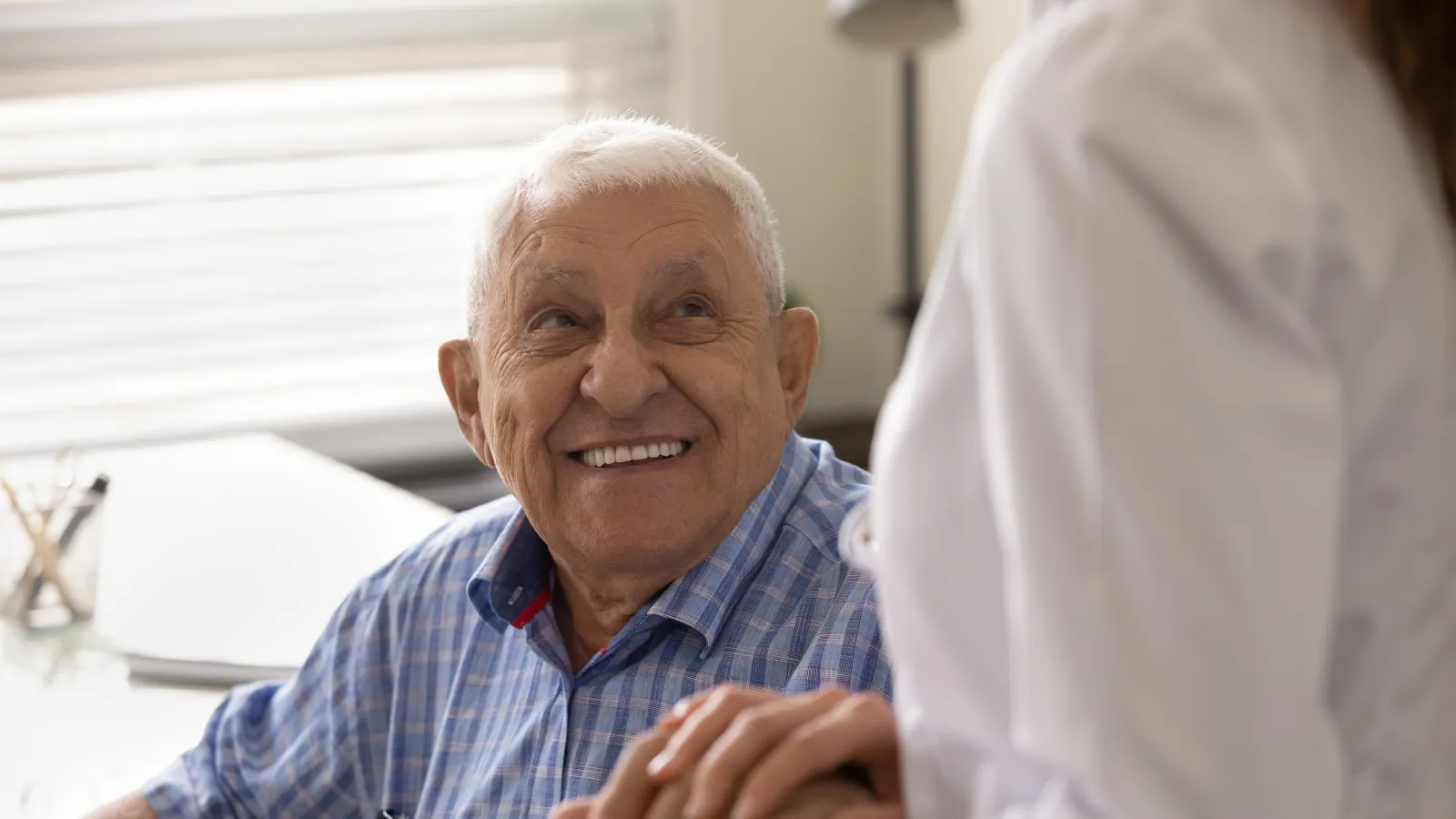 Elderly man smiling and holding hands with a healthcare professional in a bright medical office.