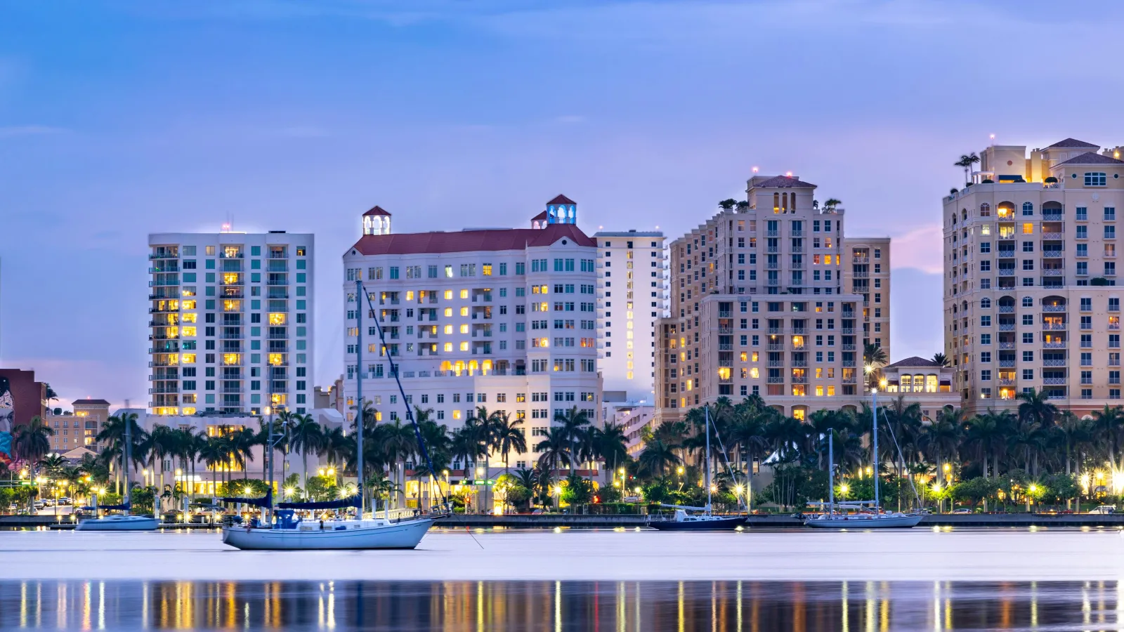 Sailboats anchored on calm water reflecting city skyline with lit buildings and palm trees at twilight.