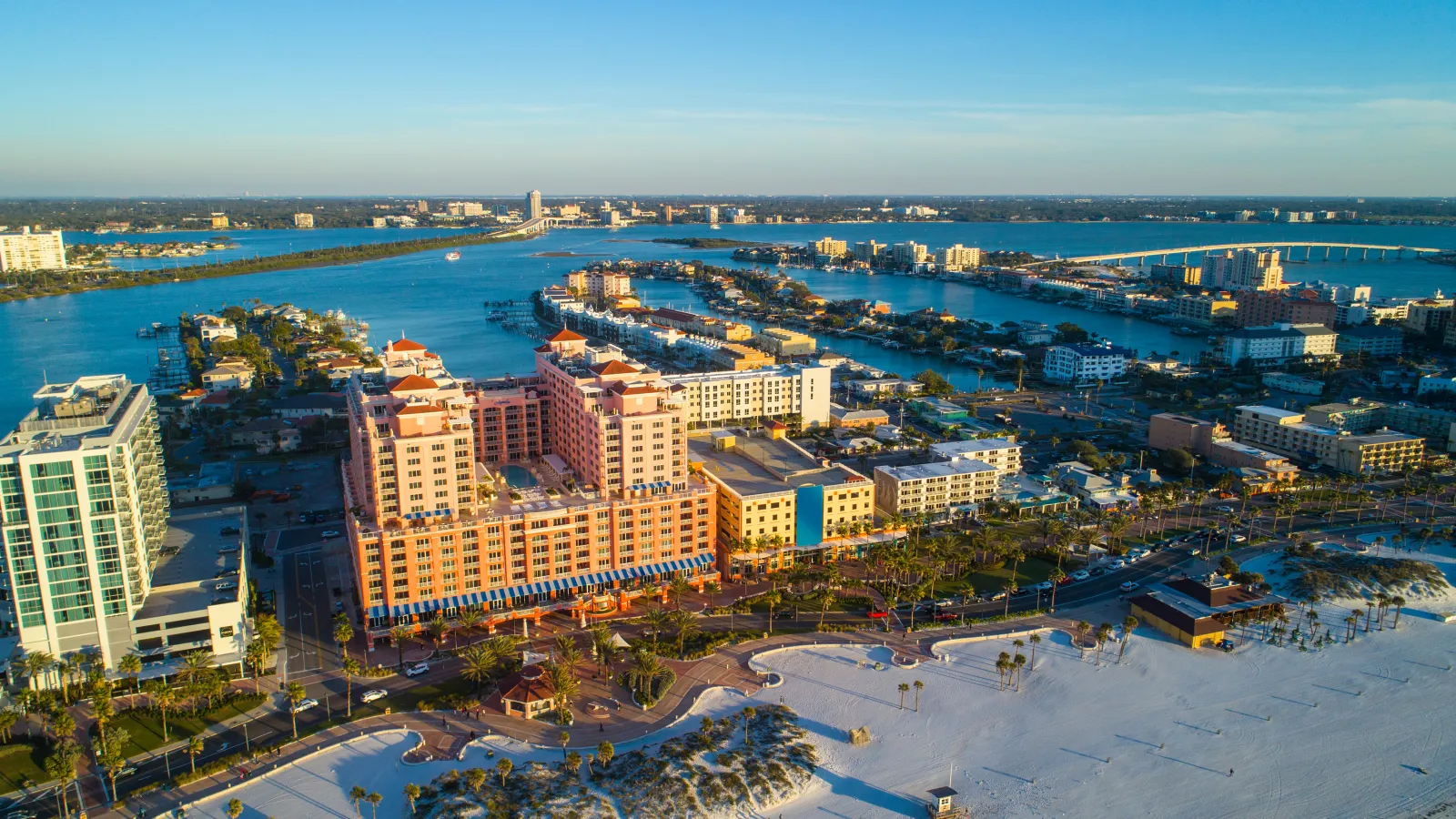 Aerial view of a coastal city with sandy beach, waterfront buildings, and a blue sky under sunlight.