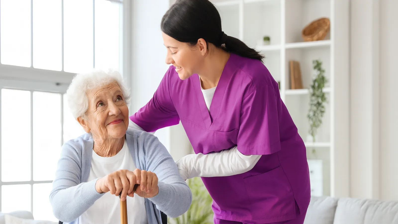 Elderly woman in a wheelchair with a caregiver in purple scrubs smiling and interacting at home.