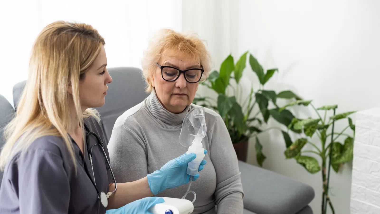 Nurse demonstrating nebulizer inhaler use to elderly woman sitting on couch in home setting.