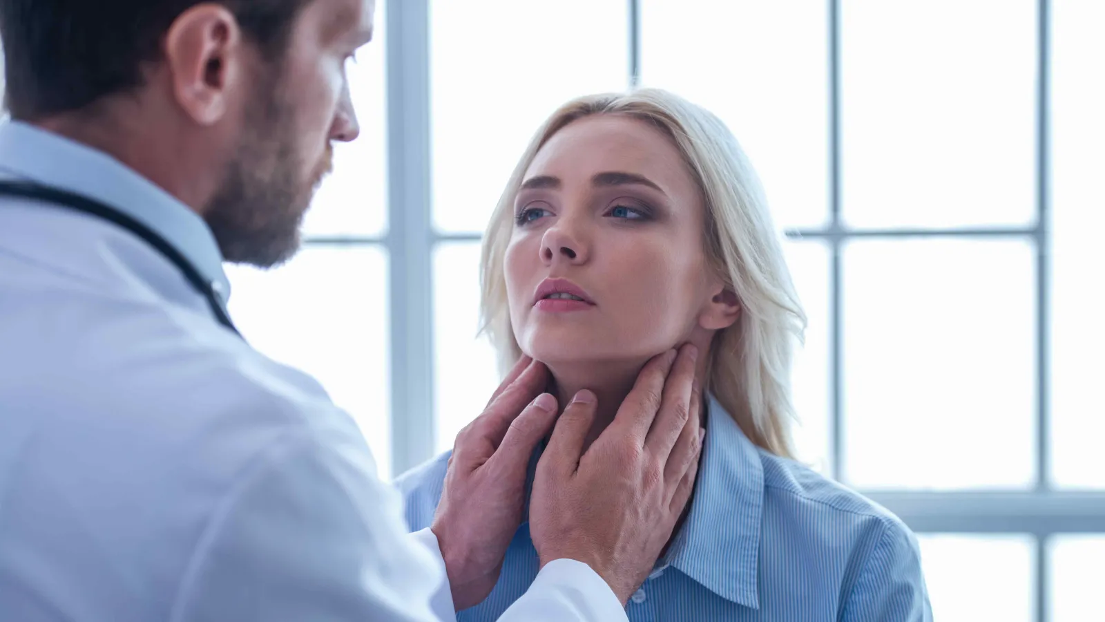 Doctor examining woman's throat in medical office with large windows and bright natural light.