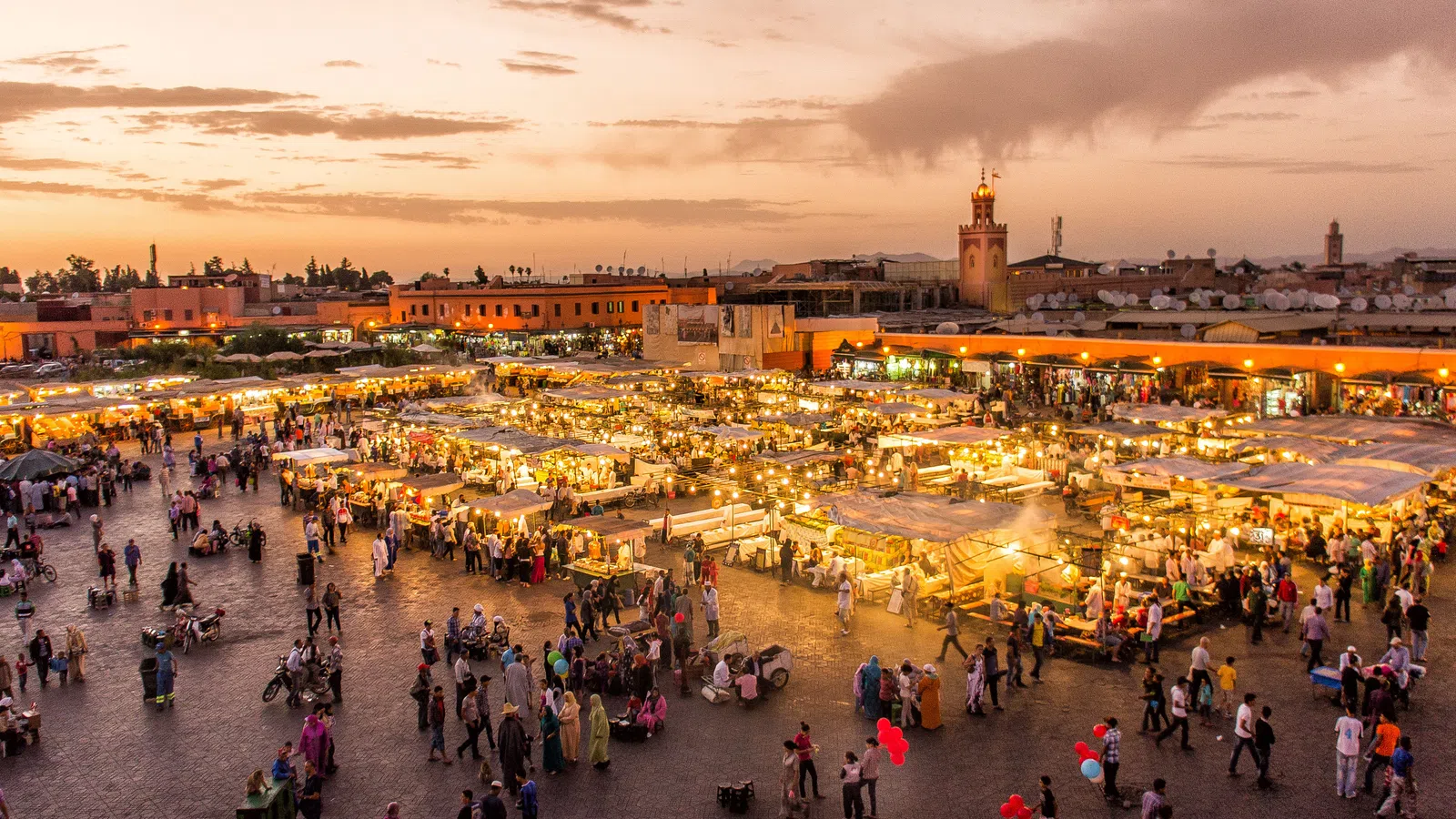 Evening aerial view of a bustling market square with illuminated stalls and crowds at sunset.