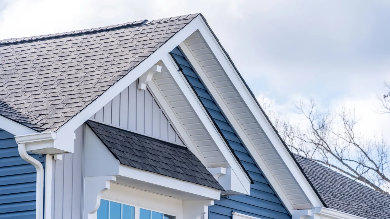 Close-up of blue house exterior with white trim, gable roof, and multiple window shades under cloudy sky.