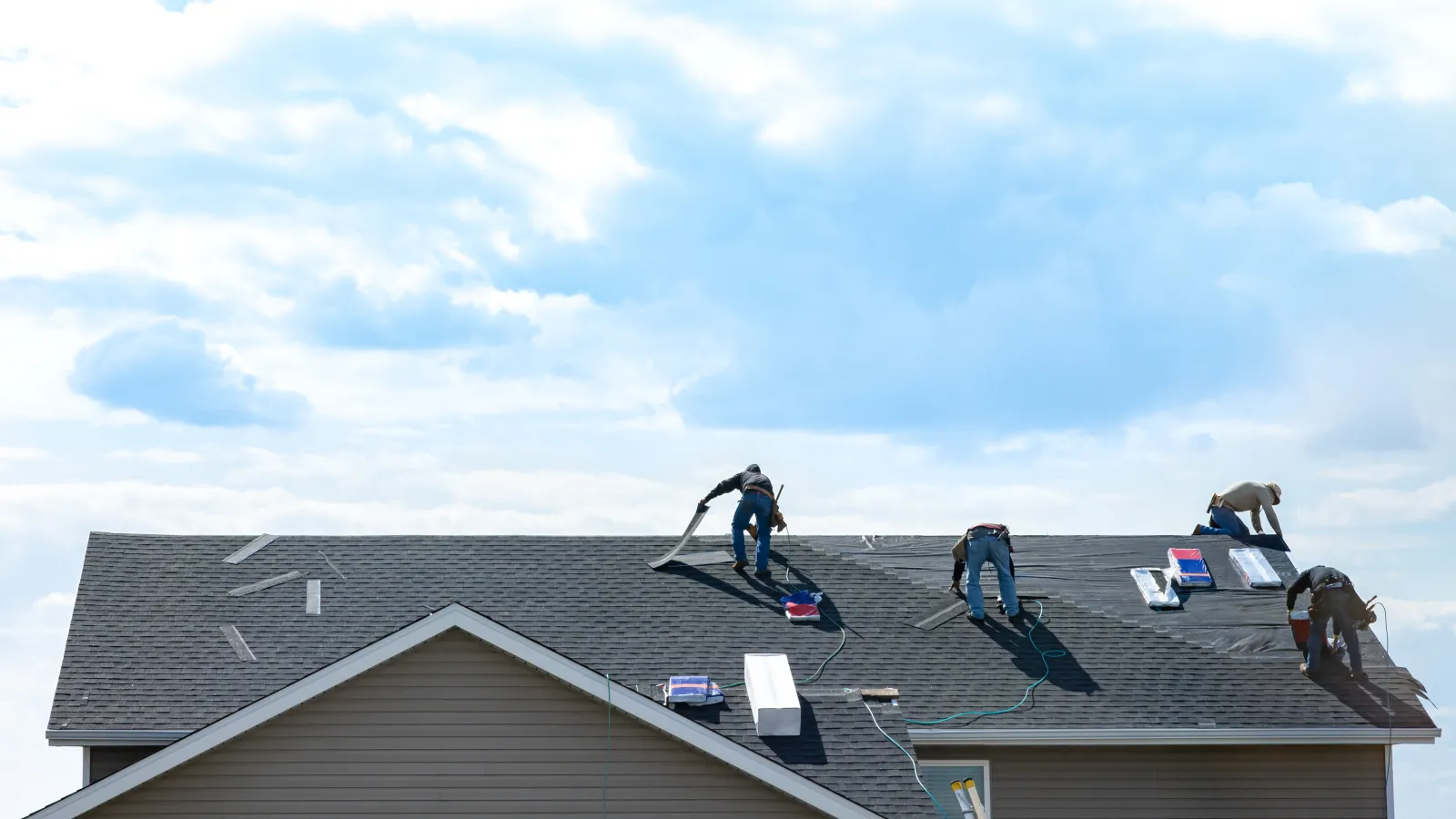 Four workers installing shingles on a residential roof under a partly cloudy blue sky.