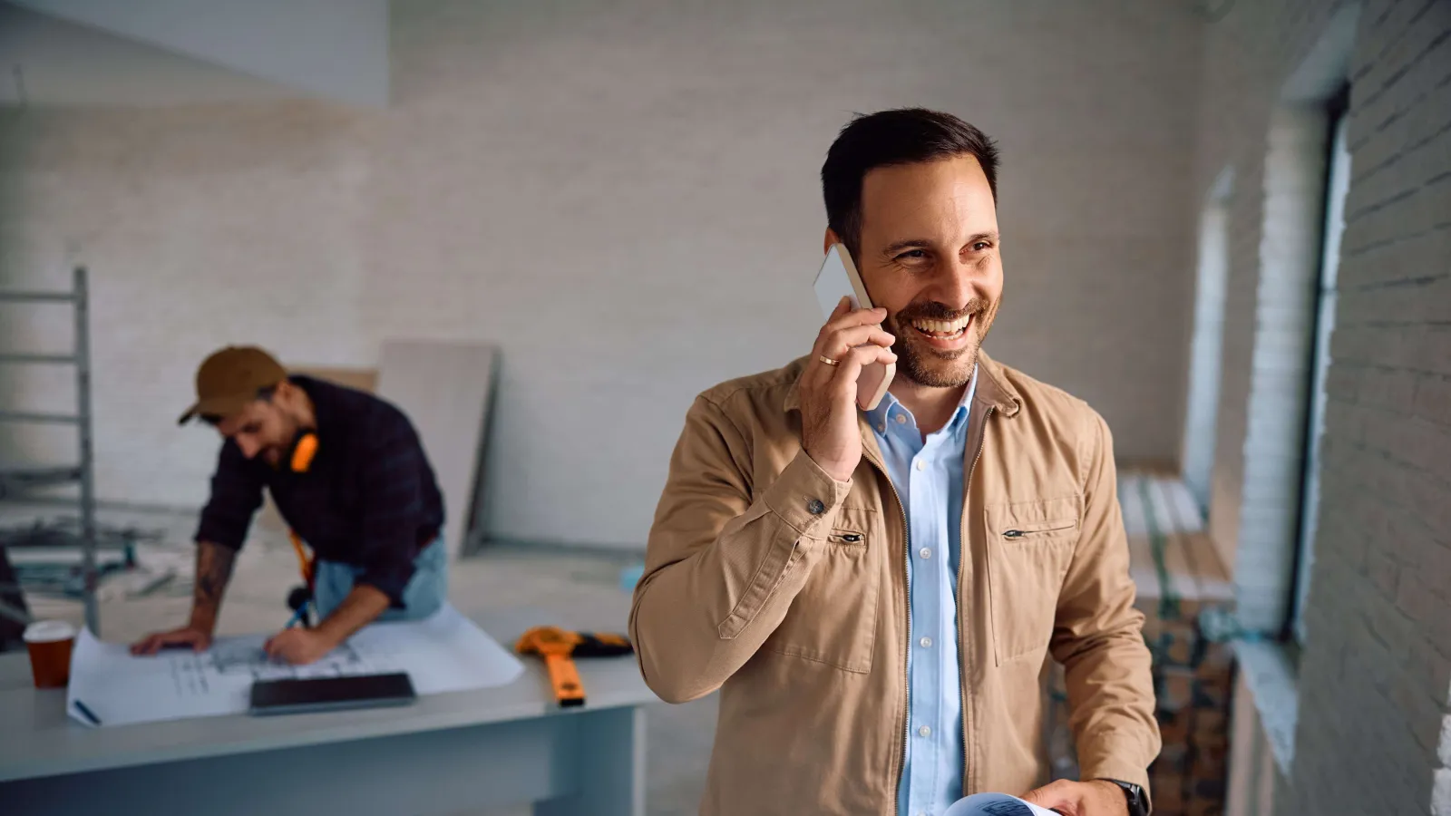 Smiling man in beige jacket talks on phone holding rolled blueprints inside construction site office.