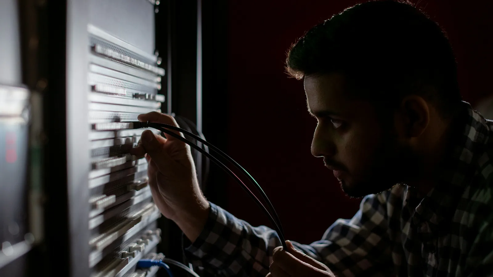 Man connecting cables to a server rack in a dimly lit data center environment focused on networking tasks.