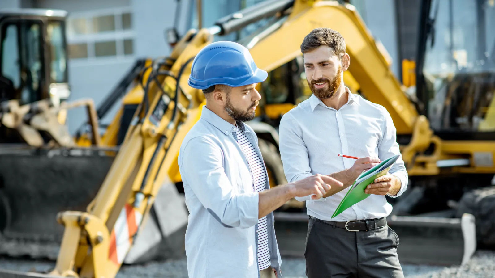 Two men discuss construction plans at a worksite with heavy machinery in the background