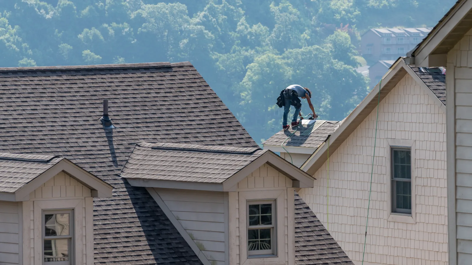 Roofer working on shingled roof of a beige house surrounded by green forested hills in the background.