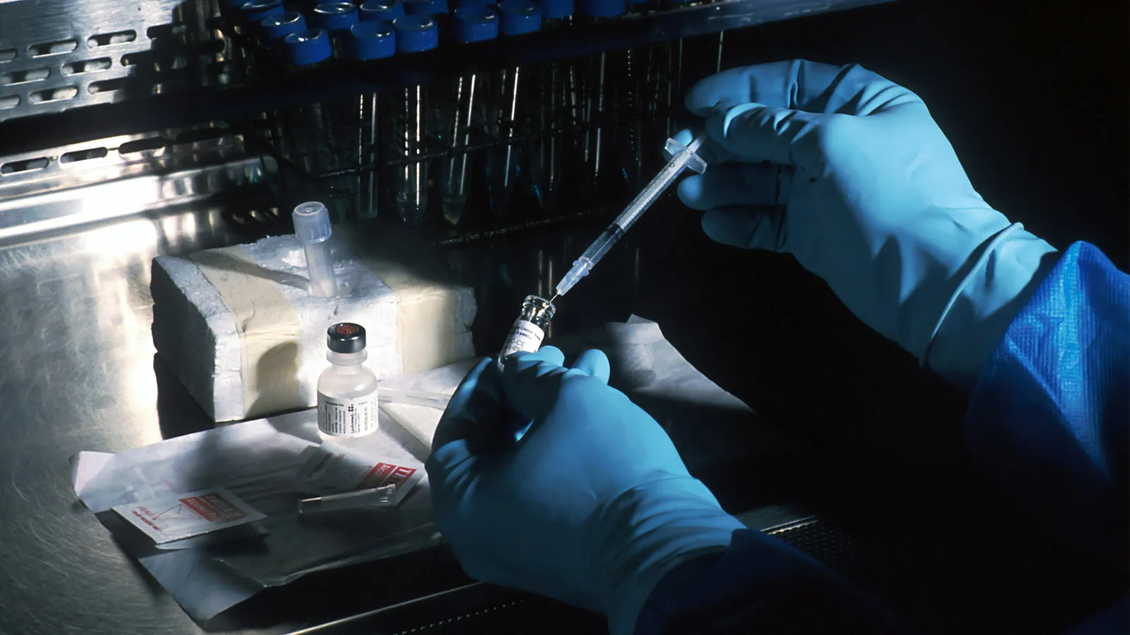 Gloved hands drawing liquid from a vial with a syringe in a sterile laboratory setting under low light.