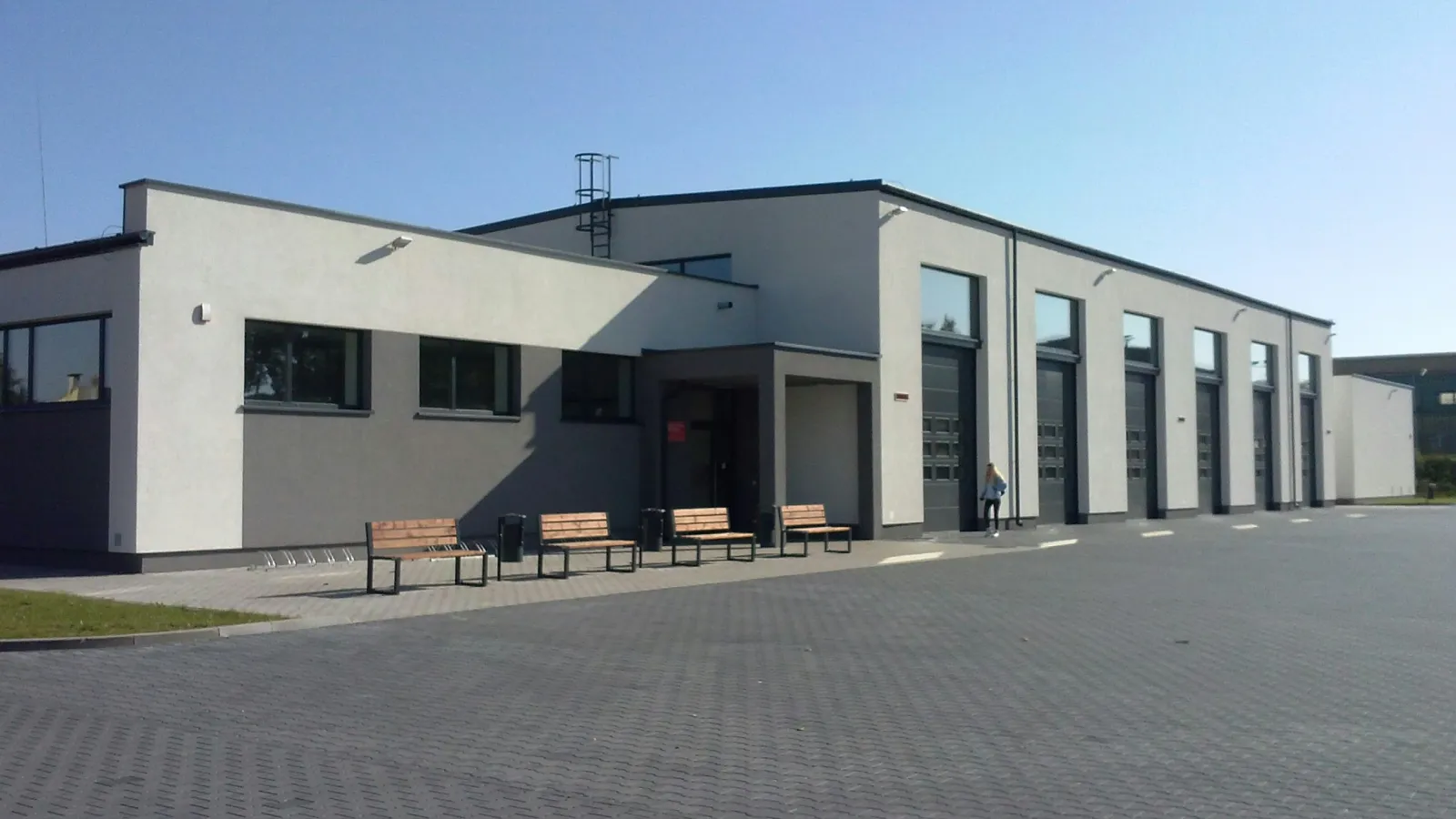 Modern white and gray industrial building with benches outside and large garage doors under clear blue sky.