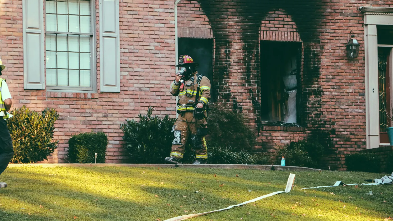 Firefighter in full gear walks outside a charred brick house after a fire incident during daylight.