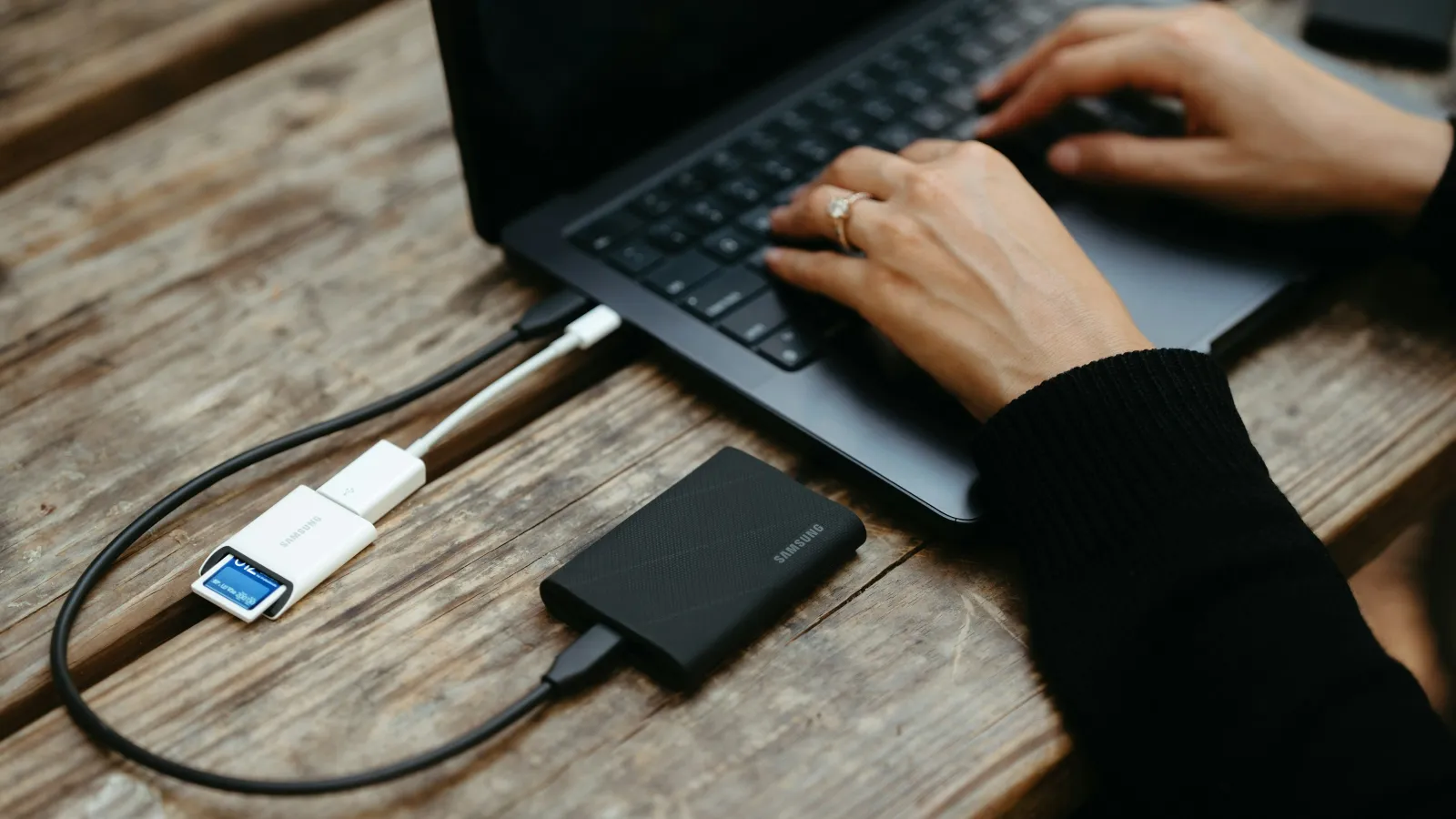 Person working on laptop connected to external Samsung SSD and SD card reader on wooden table.