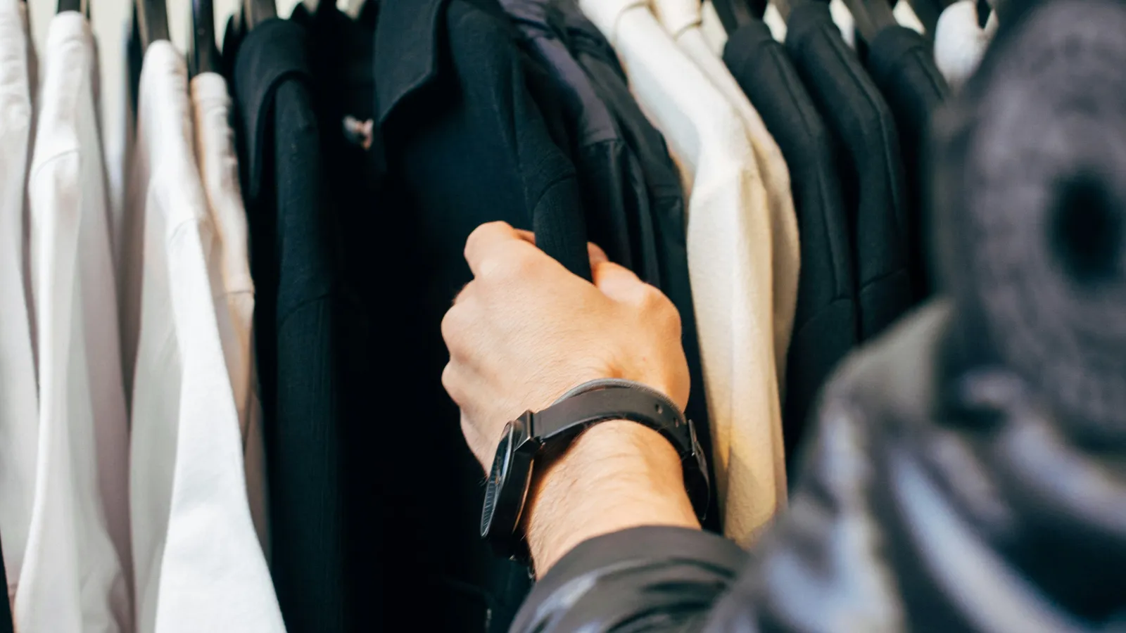 Person browsing black and white clothes hanging on a rack in a store with focus on hand and watch.