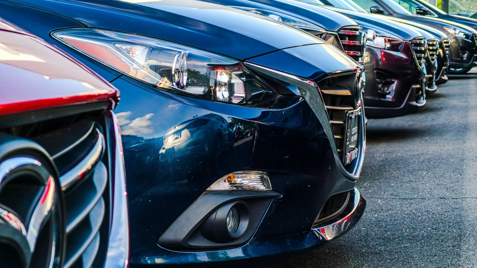 Line of parked cars with shiny front grilles and headlights in a dealership lot on asphalt pavement.