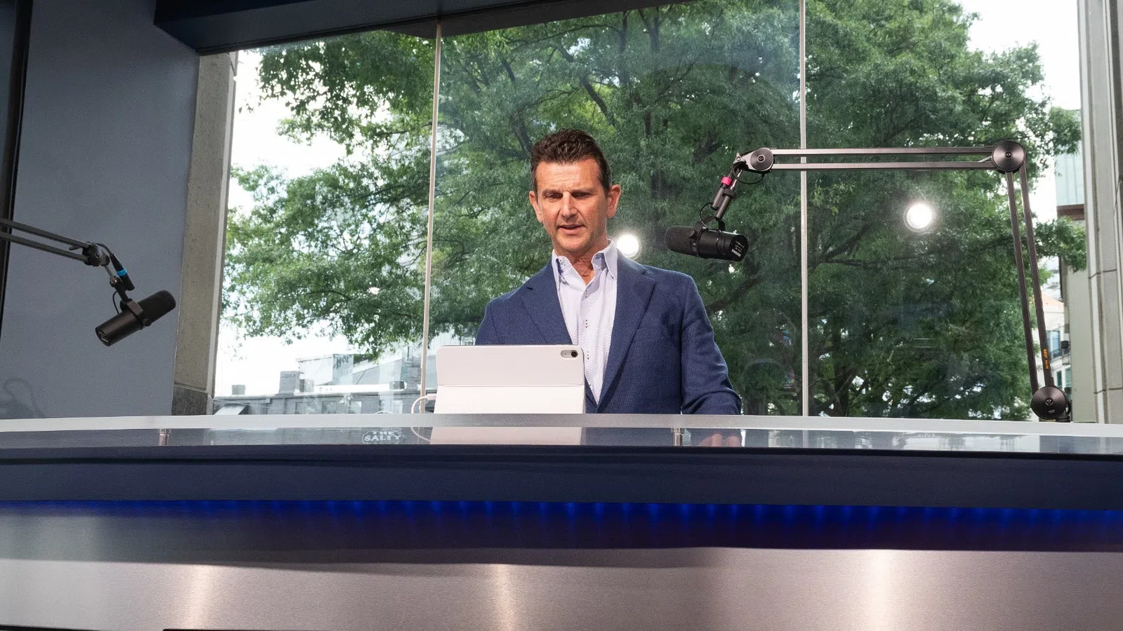 Businessman in studio presenting behind glass desk with cityscape screens and microphones in background