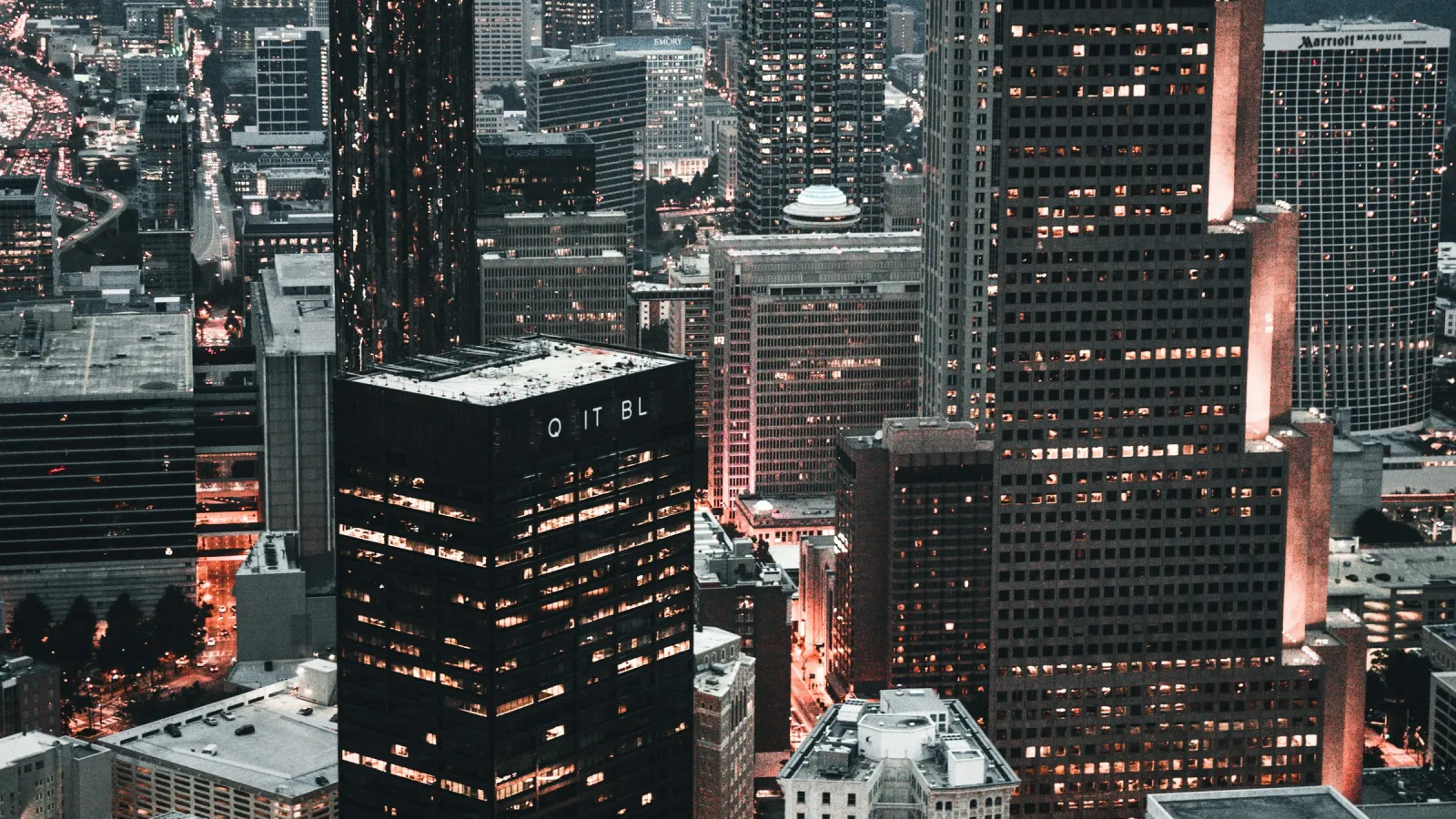 Aerial view of a city skyline at dusk with illuminated skyscrapers and busy streets glowing with orange lights.