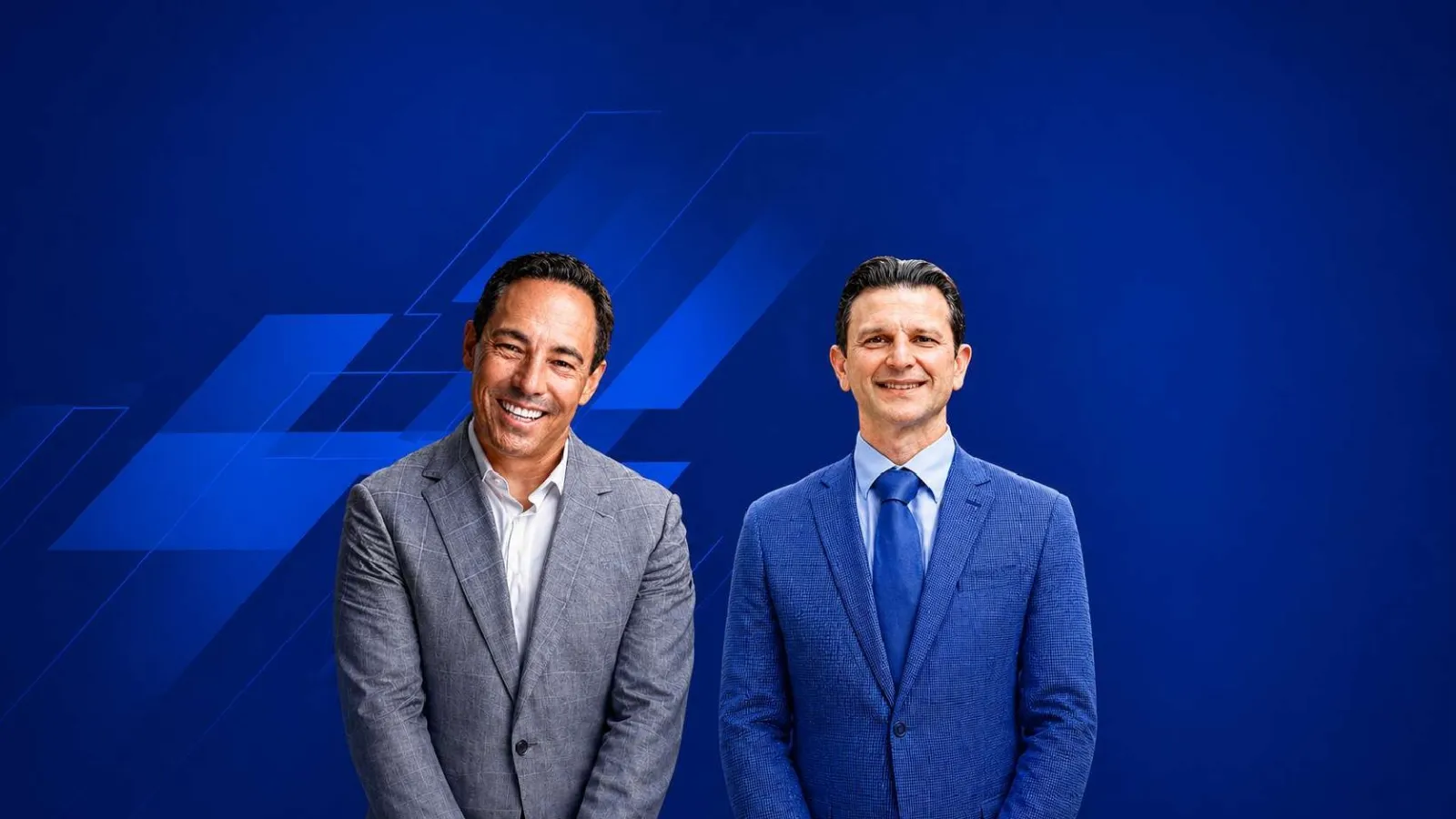 Two smiling men in business suits standing against a blue geometric background in a professional setting