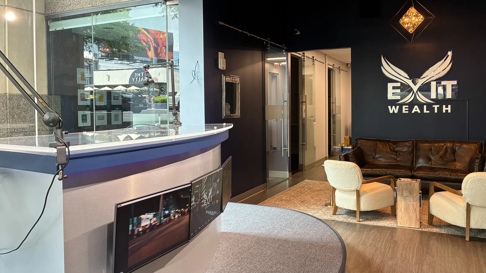 Modern office lobby with curved reception desk, monitors, leather sofa, chairs, and Exit Wealth logo on dark wall.