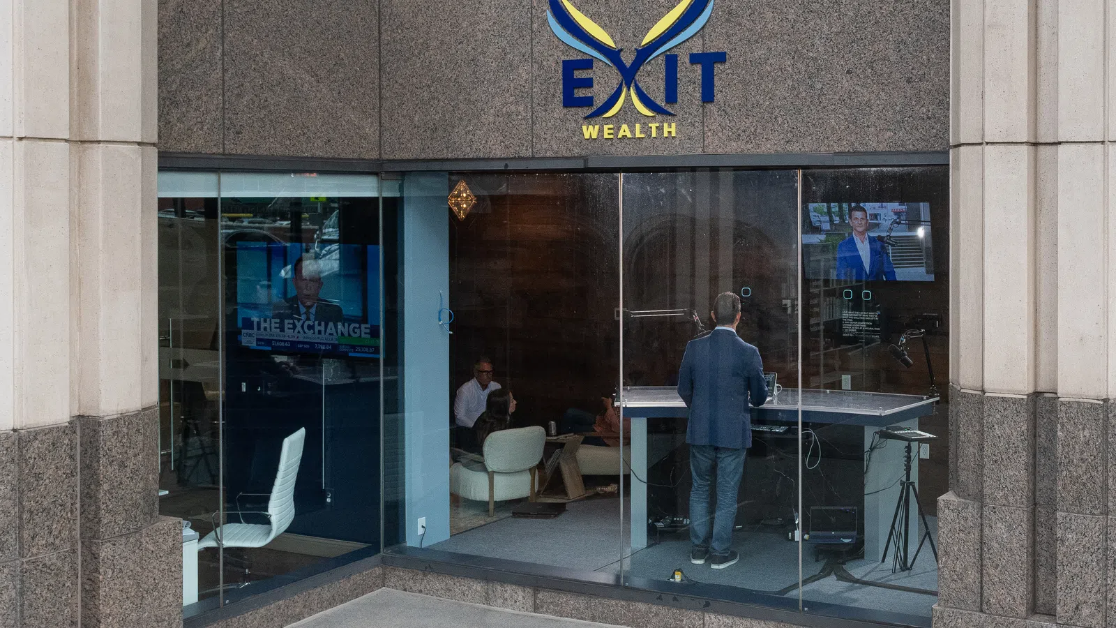 Exterior view of Exit Wealth office with glass walls showing people inside and a man standing at a desk with monitors.