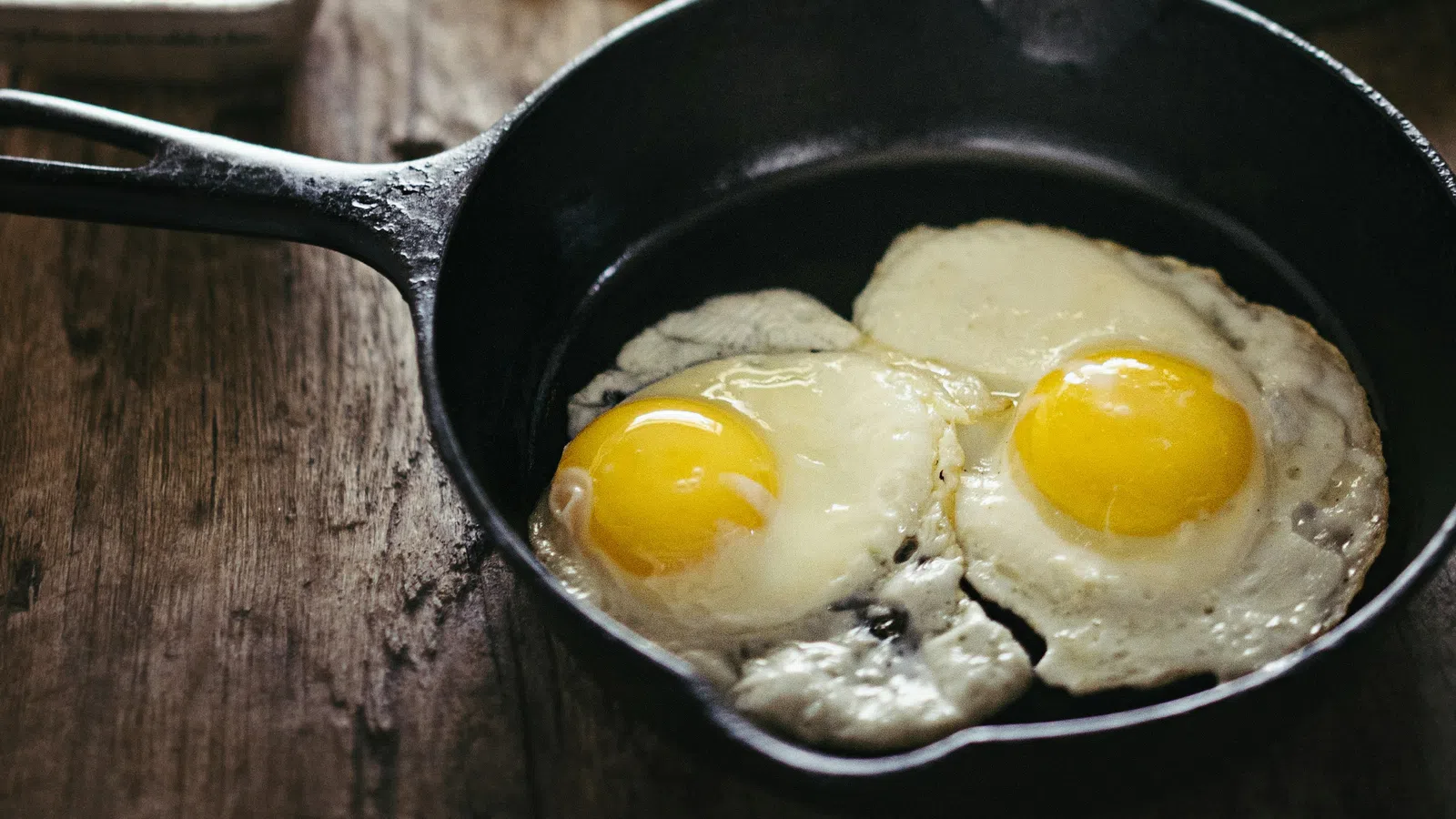 Two sunny-side-up eggs in a black cast iron skillet on a rustic wooden table with breakfast sides blurred.