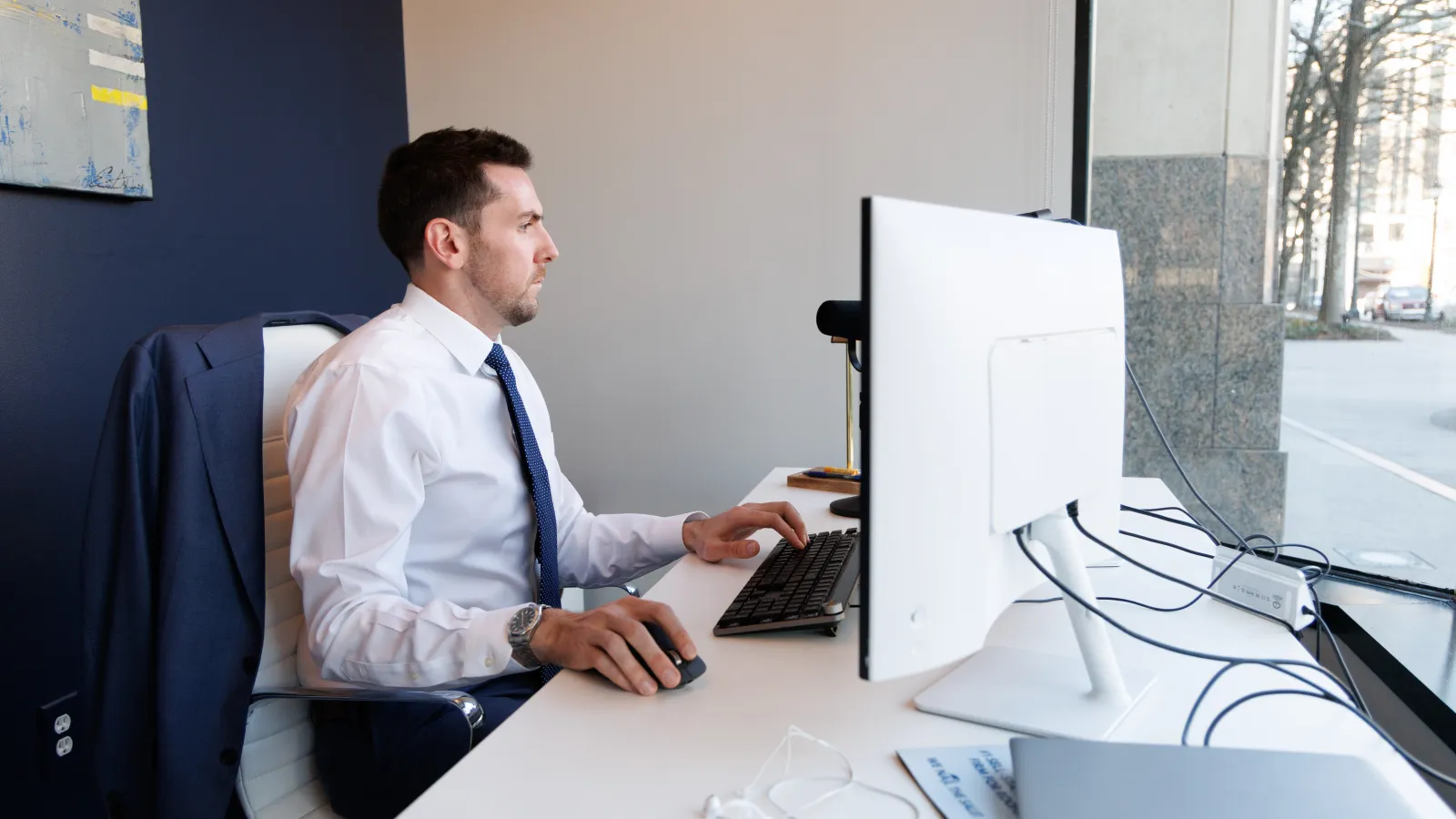 Man in business attire working at a computer desk near a window in a modern office setting.