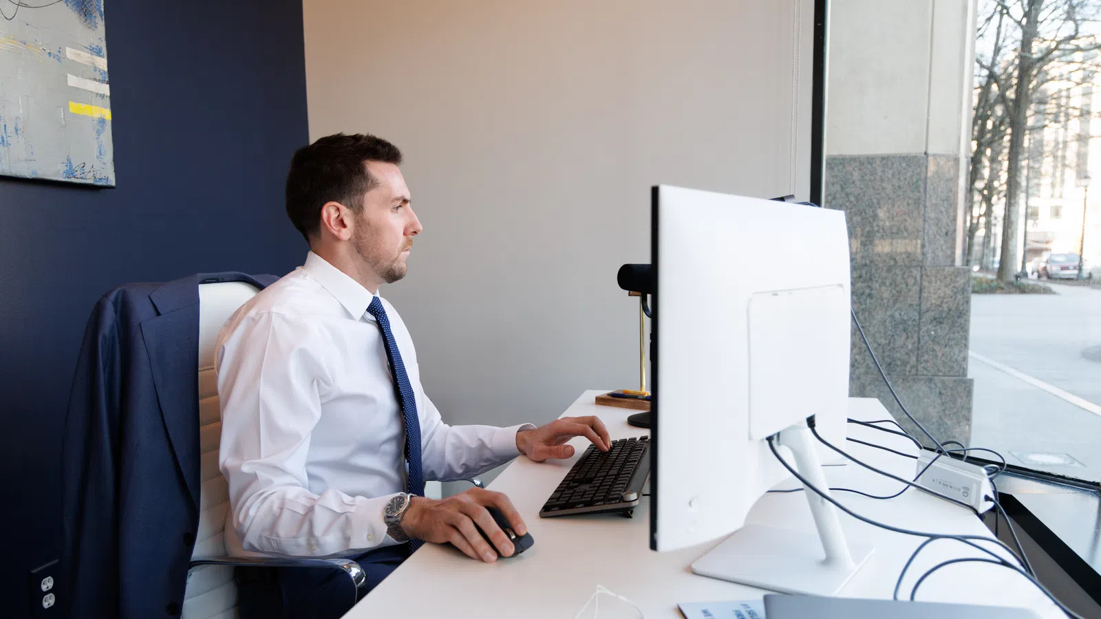Man in business attire working at a computer desk near a window in a modern office setting.
