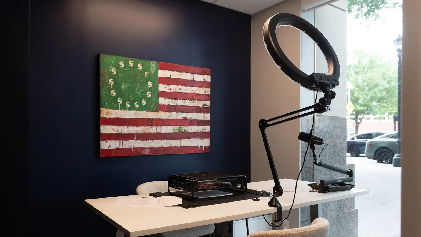 Modern office corner with white desk, ring light, American flag artwork with dollar signs, and large window view.