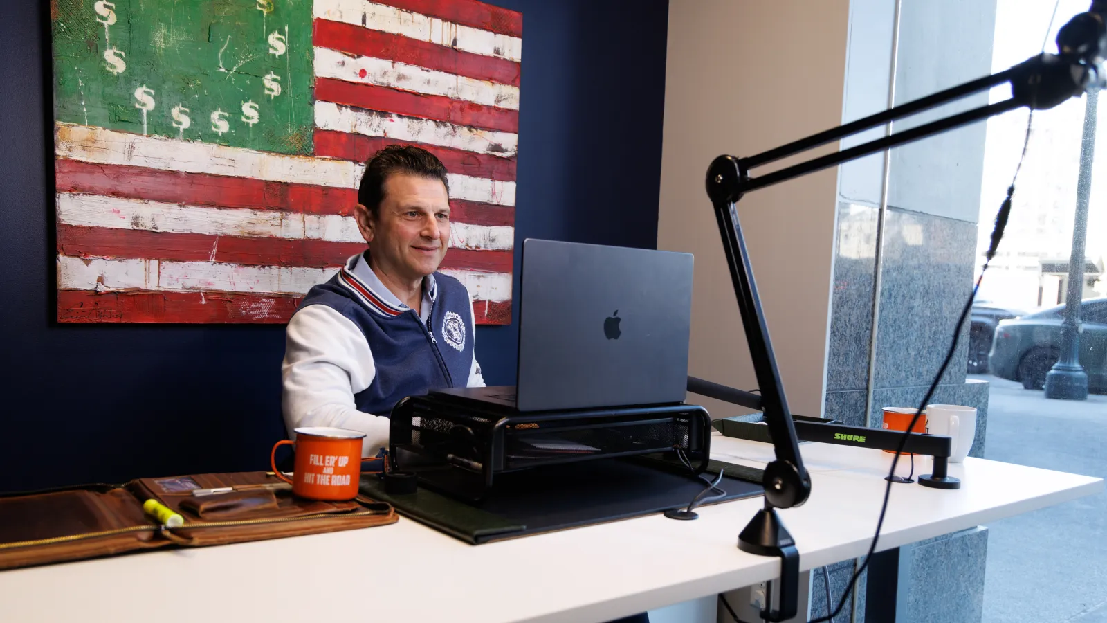 Man working on a laptop at a white desk with a microphone arm and a dollar-themed American flag painting behind him.